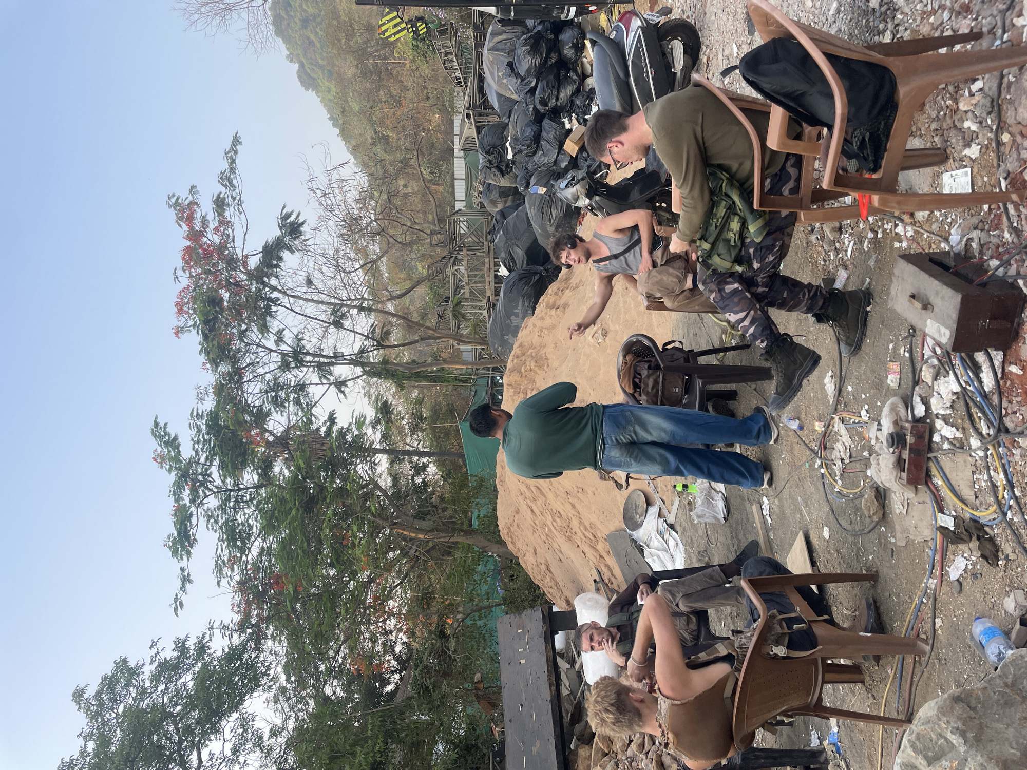 The image depicts an outdoor scene in what appears to be a cluttered or semi-constructed area, possibly within Mira-Bhayander, India, though the location is not explicitly evident.
There are six individuals present. Two are seated in plastic chairs, with one person appearing to adjust or examine something within a bag. Another person stands beside a sand pile, their back to the camera, wearing a green shirt and jeans. A fourth person is sitting in a plastic chair as well. A pile of large black bags and other debris are in the background. Electrical wires, cables, and equipment are scattered on the ground. The environment contains evidence of construction or renovation with exposed wires.