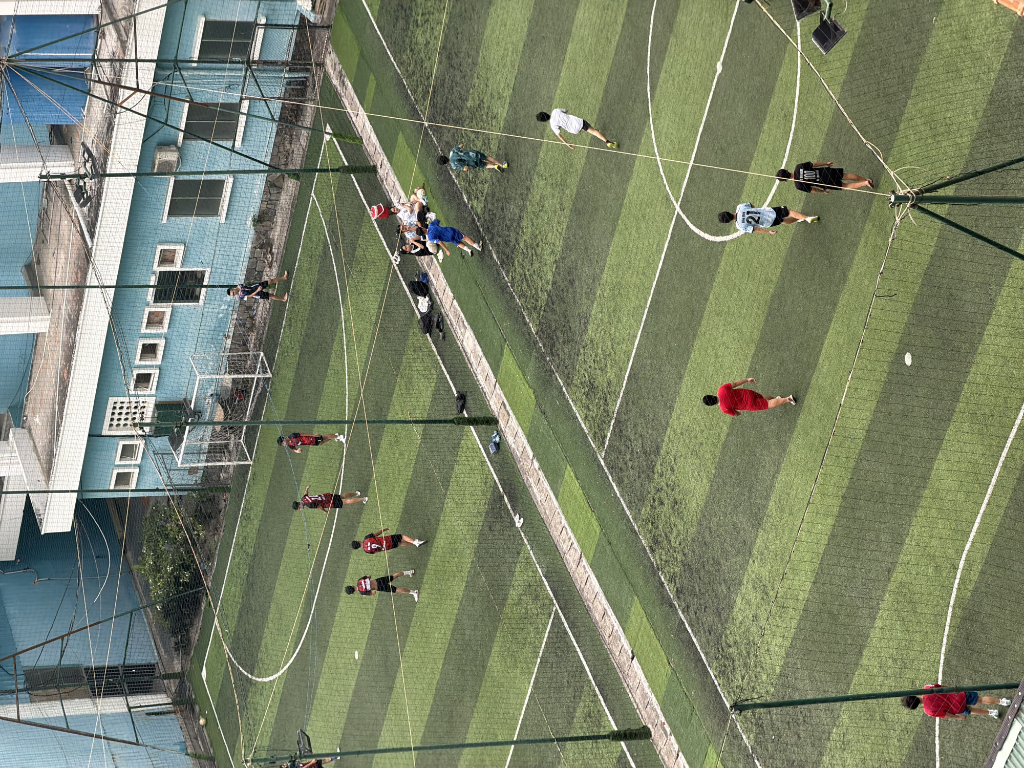 A soccer field, situated in or near Bac Nha Trang Ward, Vietnam, is depicted from an elevated perspective. Multiple individuals, identified by their athletic attire, are dispersed across the field, which is marked by green and dark green stripes with white lines defining boundaries. A building with a blue facade is located near the soccer field, potentially indicating urban development. The scene suggests an active soccer game or training session. The angle indicates that the photo was shot from a high vantage point. The field appears to be enclosed by netting.