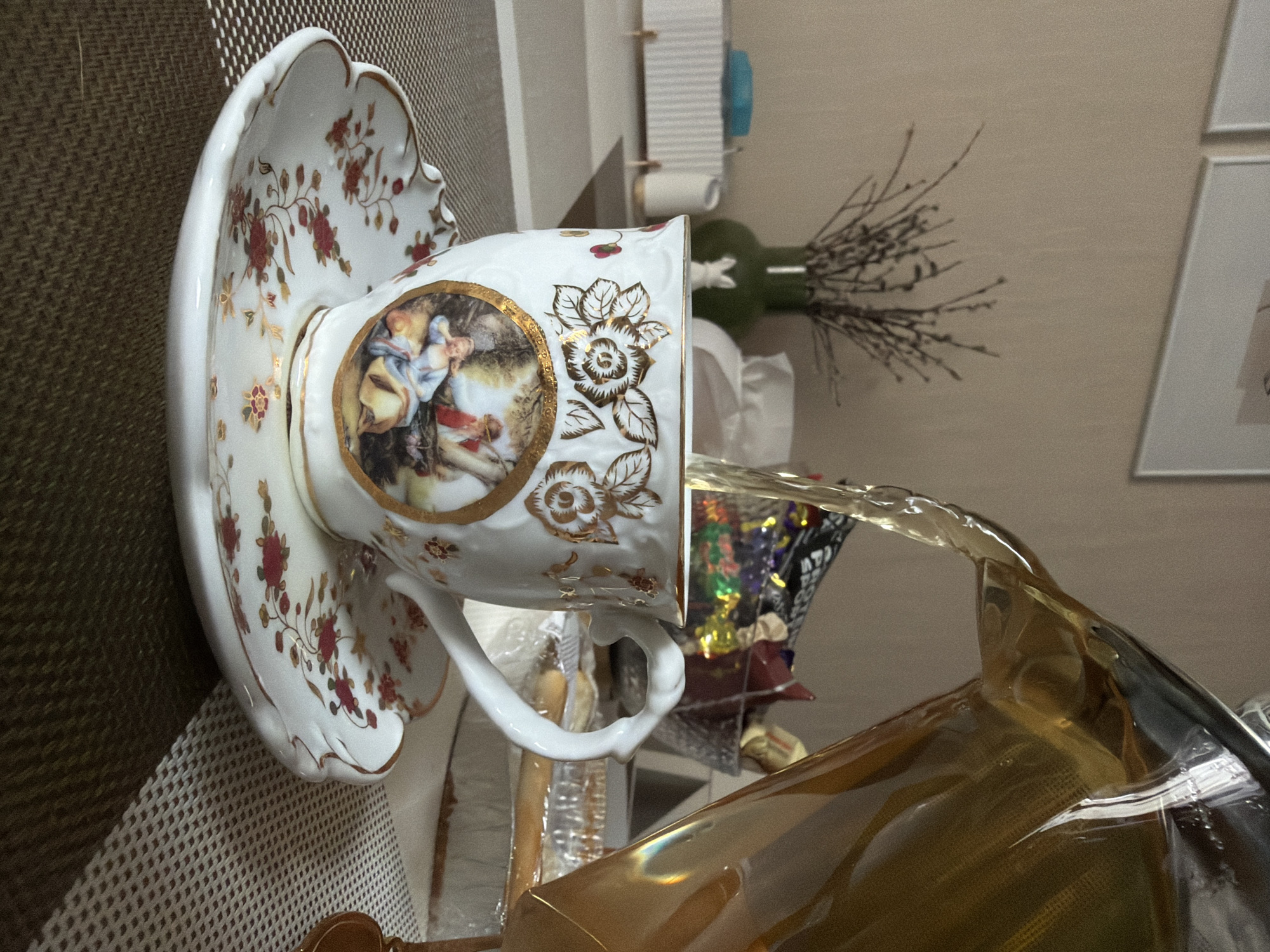The image displays a finely detailed white porcelain teacup and its matching saucer, positioned on a textured, light brown placemat. Both the cup and saucer are elaborately decorated with red floral motifs and delicate golden vine and leaf patterns. The teacup prominently features a circular, gold-framed medallion with a classical painting, depicting figures in flowing garments.
An amber-colored liquid, likely tea or a similar beverage, is being poured from a clear glass pitcher into the teacup, captured mid-stream. The pitcher is partially visible towards the lower right.
In the background, a dark green vase holds an arrangement of dried, slender branches. Behind this, a light-colored wall features two white-framed pictures. Further details in the background include a wall-mounted holder with a roll of paper and some colorful, wrapped items, possibly confections, bearing indistinct text on their packaging. The scene is set indoors, suggesting a domestic environment, possibly in Penza, Russia. The image focuses on the elegant act of pouring tea, highlighting the ornate teaware.