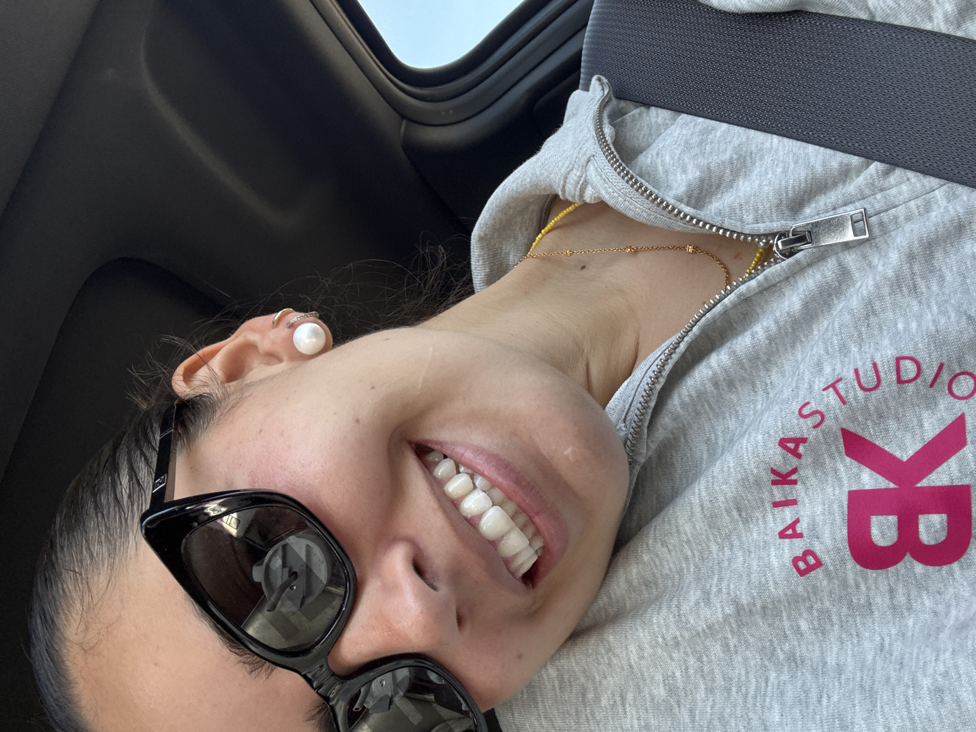 A woman wearing black sunglasses and a grey Baika Studio hoodie is in a car. She's wearing a gold necklace and a pearl earring, and she is smiling. The location is not discernible.