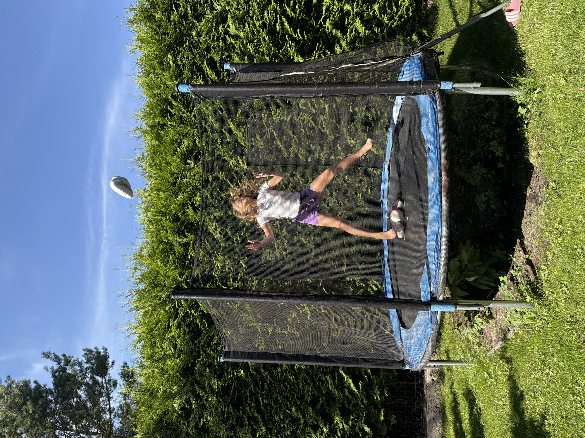 In Baloži, Latvia, a young girl with blonde, curly hair is captured mid-air on a trampoline in a backyard. She is wearing a white t-shirt and purple shorts, with her arms outstretched as she jumps. A white object, possibly a ball, is in the air above her. The trampoline is surrounded by a black safety net and is situated on a grassy lawn with a dense green hedge forming the background. The sky is clear and blue, indicating a sunny day. Pink flip-flops are visible on the grass near the trampoline.