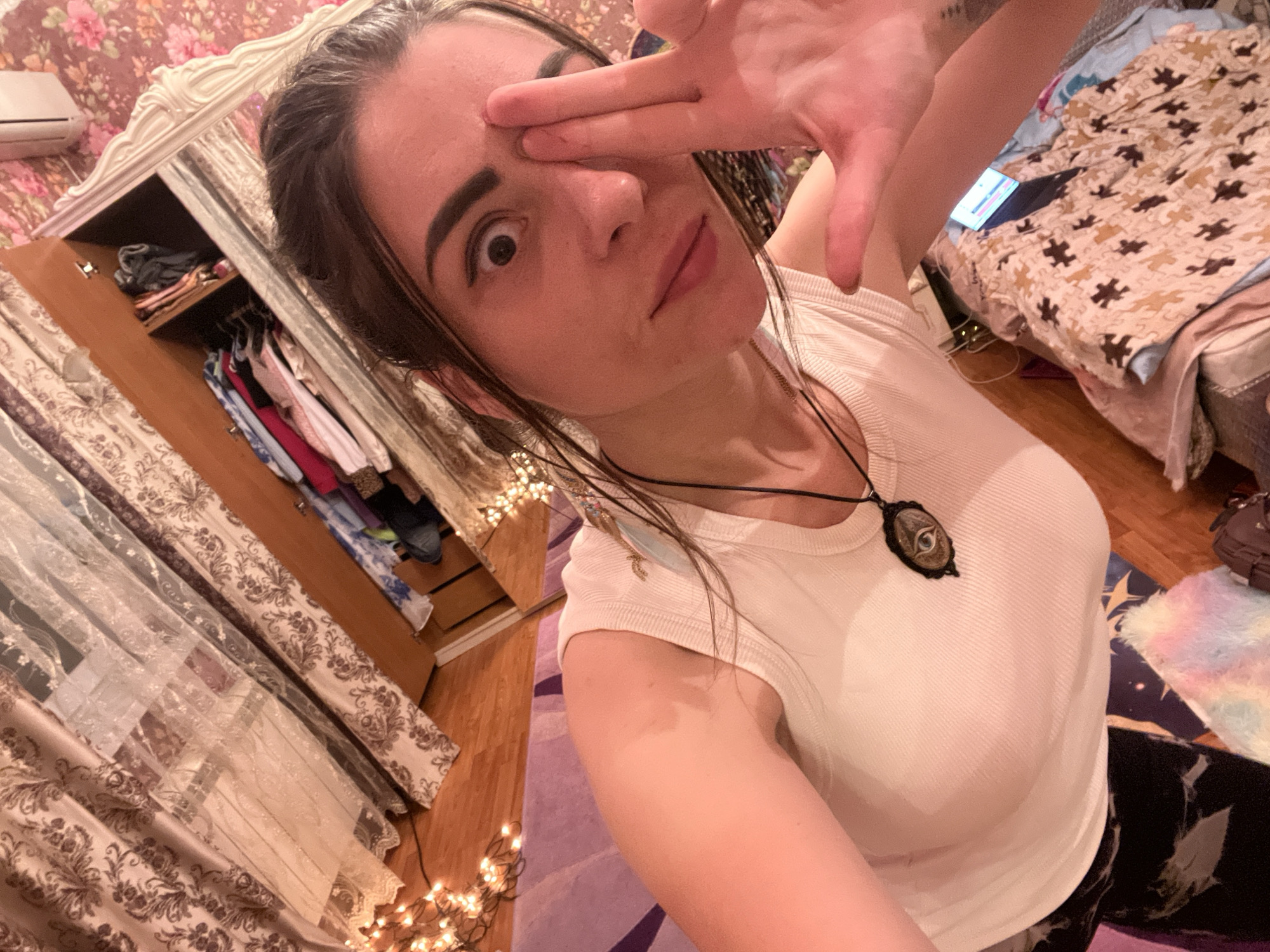 A young woman is taking a selfie in what appears to be a bedroom. She is wearing a white tank top and a necklace with an eye pendant. Her right hand is raised to her forehead, with her fingers spread out and her index finger covering her eye. Her left eye is wide open, and she has a surprised expression on her face.
The background is cluttered with various objects. To the left, there is an open wardrobe with clothes hanging inside. In the center, there is a bed with a blanket that has a puzzle-piece pattern. There is also a laptop on the bed and a small air conditioning unit on the wall. Fairy lights are strung around the room, adding a warm glow. The room appears to be decorated with floral wallpaper.
The lighting suggests it might be nighttime or indoors with artificial lighting. The overall mood seems playful and candid. There is no visible text in the image. The location context provided is Krasnoe-na-Volge, Russia, but there are no specific visual cues in the image to confirm this.