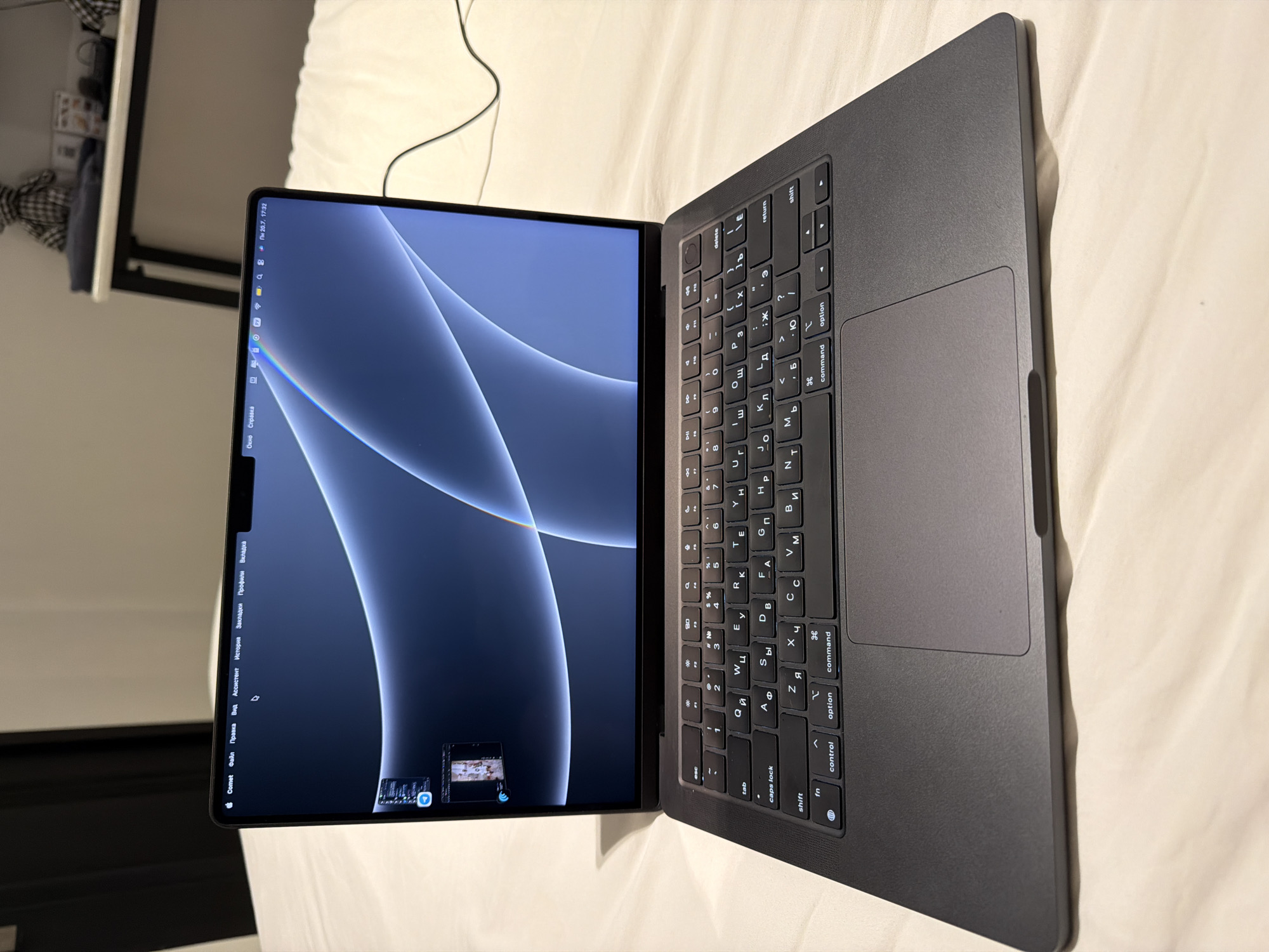 A dark gray MacBook is open and lying on a light-colored, textured surface, likely a bed. The laptop screen displays a bluish-gray abstract wallpaper with flowing curved lines. The keyboard is visible and appears to be in Russian. Several icons are visible in the menu bar at the top of the screen, including a Wi-Fi symbol, battery indicator, and clock showing "20.7. 17:32". To the left of the screen, there are two small thumbnail windows displayed, one above the other. The scene is indoors, with a dark doorway visible in the background on the left. The overall impression is of a personal workspace or leisure setting. The image is captured from a slightly elevated, forward-facing perspective. There is no discernible connection to Ban Phonsavang Neua, Laos.