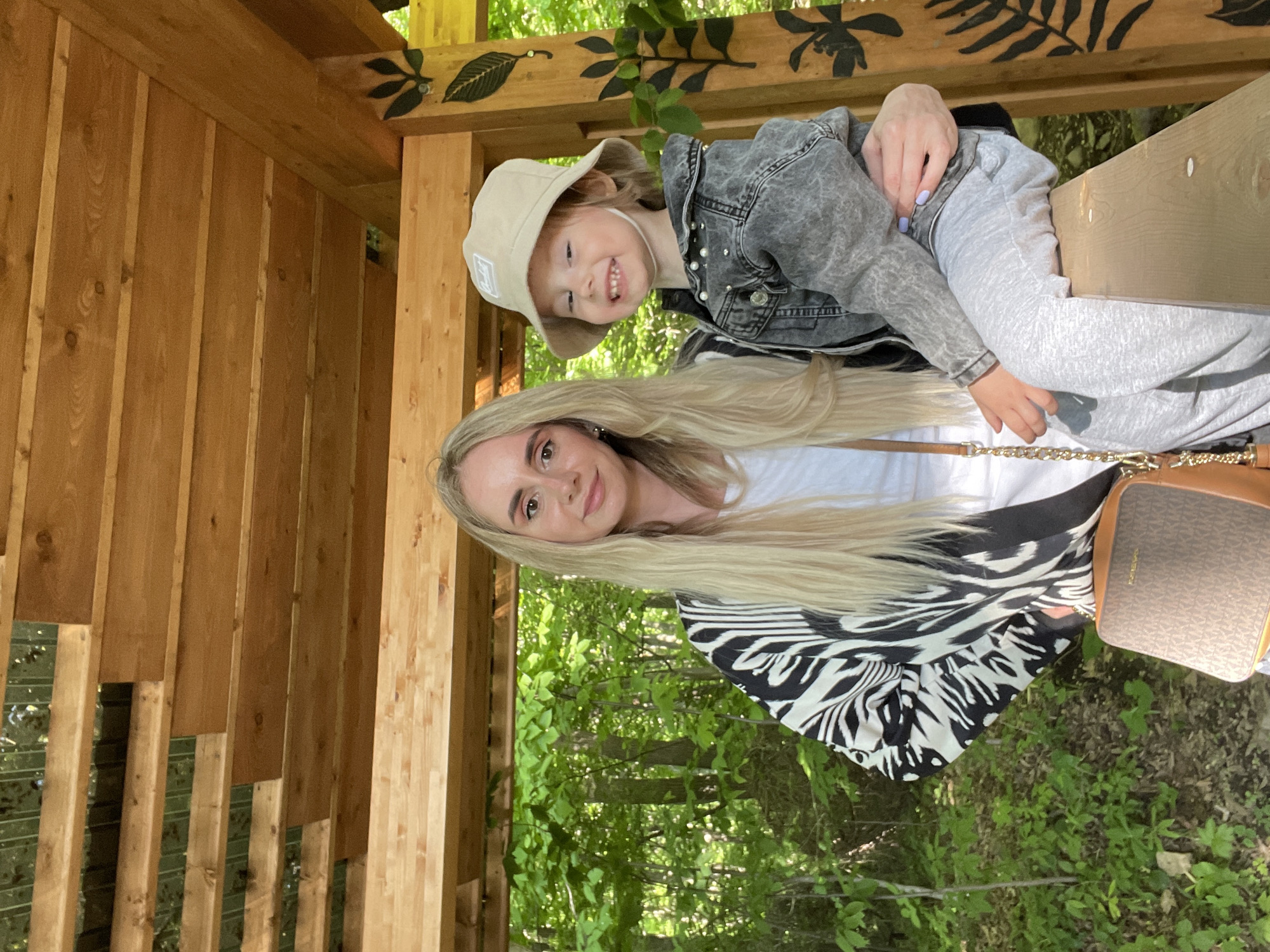 A woman with long blonde hair, wearing a white t-shirt and a zebra-print jacket, is holding a young child with a bucket hat and grey denim jacket. The child is smiling and looking at the camera. They are standing in a wooden structure with green foliage in the background. It is not possible to determine if the image was taken in Vladivostok, Russia.