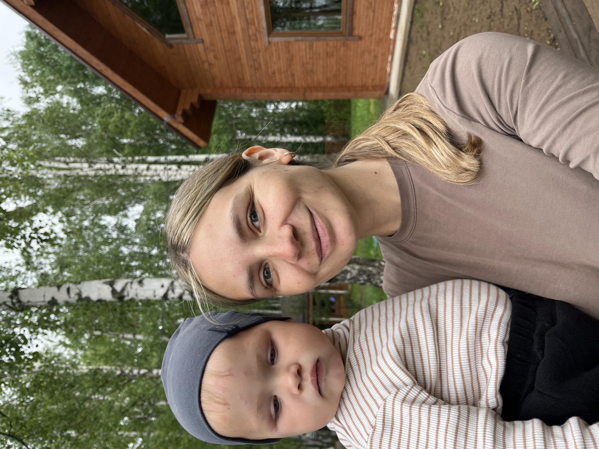 A woman and a baby are posing for a selfie outdoors. The woman has blonde hair, a fair complexion, and is wearing a brown t-shirt. The baby is wearing a gray hat and a striped shirt with dark-colored bottoms. The background includes trees, a wooden house, and birch trees with white bark and black markings. Based on the location information, the scene is likely in a residential or recreational area in the city of Chaikovsky, Russia.