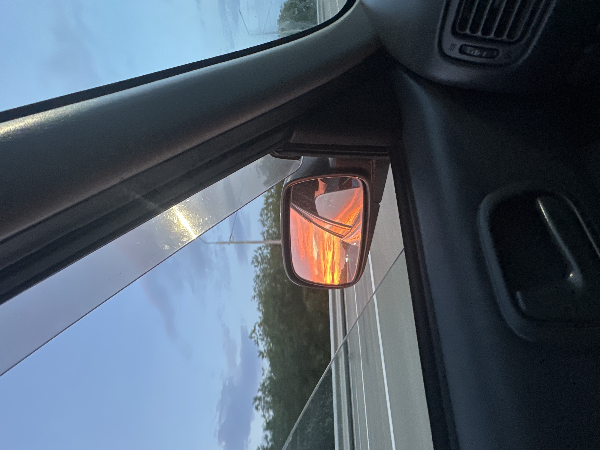 The image captures a view from inside a car, looking out the passenger window and into the side mirror. The side mirror reflects a vibrant sunset, with intense oranges and reds painting the sky. Beyond the mirror, a highway stretches out with parallel white lines, bordered by a line of dark green trees and a pale blue sky with scattered clouds. The interior of the car is visible, featuring a dark-colored door panel with a handle and an air vent. The sun is setting, casting a warm glow on the scene. The image does not provide any specific information about the city of Yelizovo, Russia.