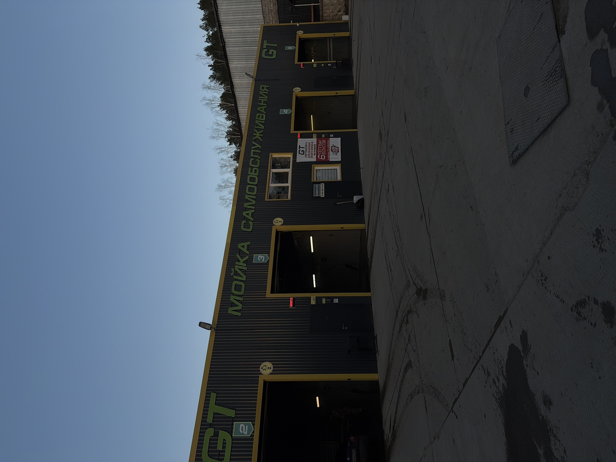 The image depicts the exterior of a car wash named "GT Self-Service" located in the city of Chaikovsky, Russia. The building is a dark, corrugated metal structure with yellow trim around the entrances to the car wash bays. Large green lettering on the building spells out "МОЙКА САМООБСЛУЖИВАНИЯ" (Self-Service Car Wash). There are several numbered bays, with at least five visible (numbered 2 to 5). Signage indicates services are available 24/7. A concrete area is in the foreground, possibly a driveway or waiting area. The sky is visible above the building, suggesting it is daytime. A line of trees is visible behind the building.