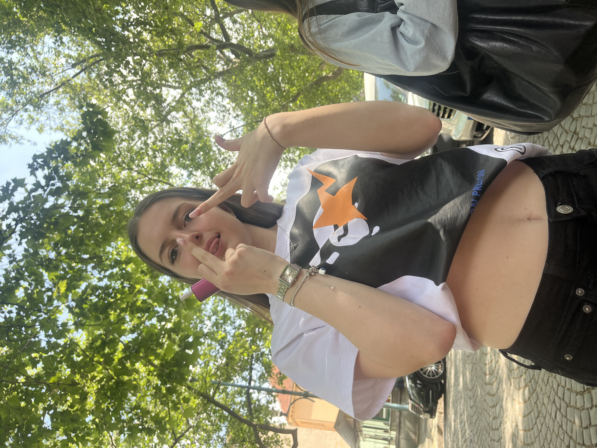 A young woman is outdoors, possibly in a park or urban environment, as evidenced by the presence of trees and paving stones. She is wearing a white t-shirt with a large black graphic design featuring an orange star. She is also wearing black pants and several bracelets on her left wrist, along with a watch. The woman is holding a pink cylindrical object in her left hand and is making a gesture with her right hand. She is sticking out her tongue. In the background, there are trees, a building with a reddish roof, and possibly a car. The image's orientation suggests it was taken with a mobile device held at an angle. It is not possible to determine the location in the city of Plovdiv, Bulgaria from the current image.