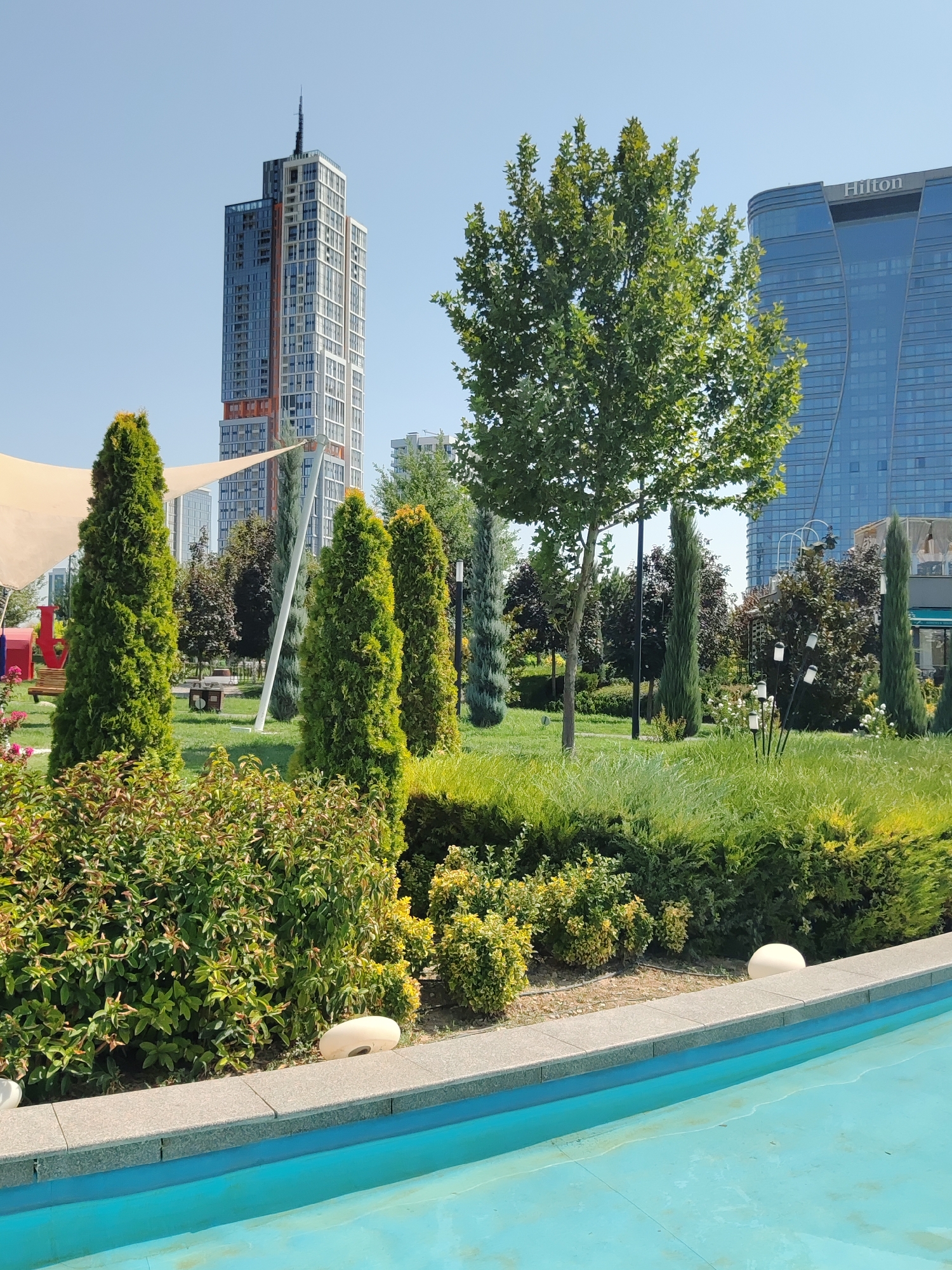 This is a photograph of a public park in Tashkent, Uzbekistan, featuring a modern cityscape in the background. In the foreground, a bright blue pool or water feature is visible, bordered by a stone edge. Behind the pool, well-maintained green bushes and hedges form a manicured landscape. Several tall, slender evergreen trees are planted in a row, with a larger deciduous tree in the middleground.
Dominating the background are two prominent buildings. To the left, a very tall, modern skyscraper with a distinctive design featuring various colored panels and a spire at its peak stands against a clear blue sky. To the right, a contemporary glass-faced building with the "Hilton" logo prominently displayed is visible, its curved facade reflecting the sky. Scattered throughout the park are benches and lighting fixtures, suggesting it is designed for public use. The overall scene depicts a blend of urban development and green recreational space, bathed in bright sunlight.