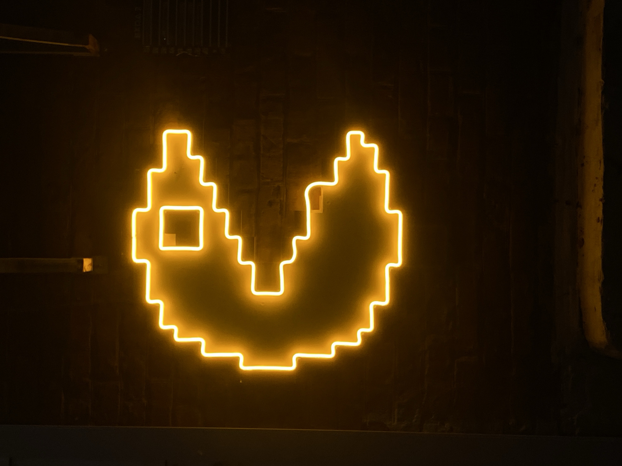 A glowing yellow neon sign depicting Pac-Man is mounted on a dark brick wall. The sign is pixelated in design. A metal grate is visible at the top of the image. To the right of the Pac-Man sign is a corroded and painted pipe. A ledge is visible at the bottom of the image. The image was taken at night or in a dimly lit location. The image shows no people, subjects or actions. There is no location information.