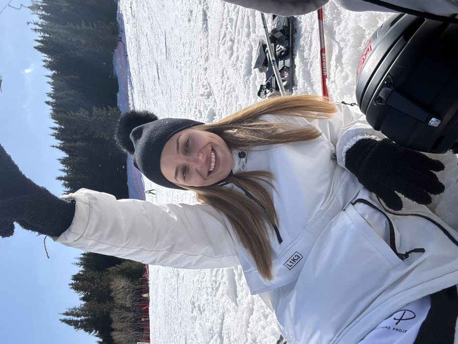 This image displays a female subject positioned on a snow-covered ski slope in Bistritsa, Bulgaria, under a clear blue sky. The woman, appearing to be a young adult with long, light brown hair, is looking directly at the camera with a wide smile, exposing her teeth and dimples. She is dressed in a white ski suit featuring black zippered accents and the text "L1K3" on the chest. She wears a black knit beanie with a black fur pom-pom and black, textured gloves. Her left arm is raised vertically in the air, while her right arm rests on the snow. To the subject's right, ski equipment is present on the snow, including ski bindings, a red ski pole with "Salomon" visible, and a partially visible black ski helmet in the lower right foreground. The background reveals an expansive, groomed snow slope, with red safety netting positioned on the left side and several indistinct figures of other skiers in the distance. A dense coniferous forest forms the horizon line beneath the clear sky.