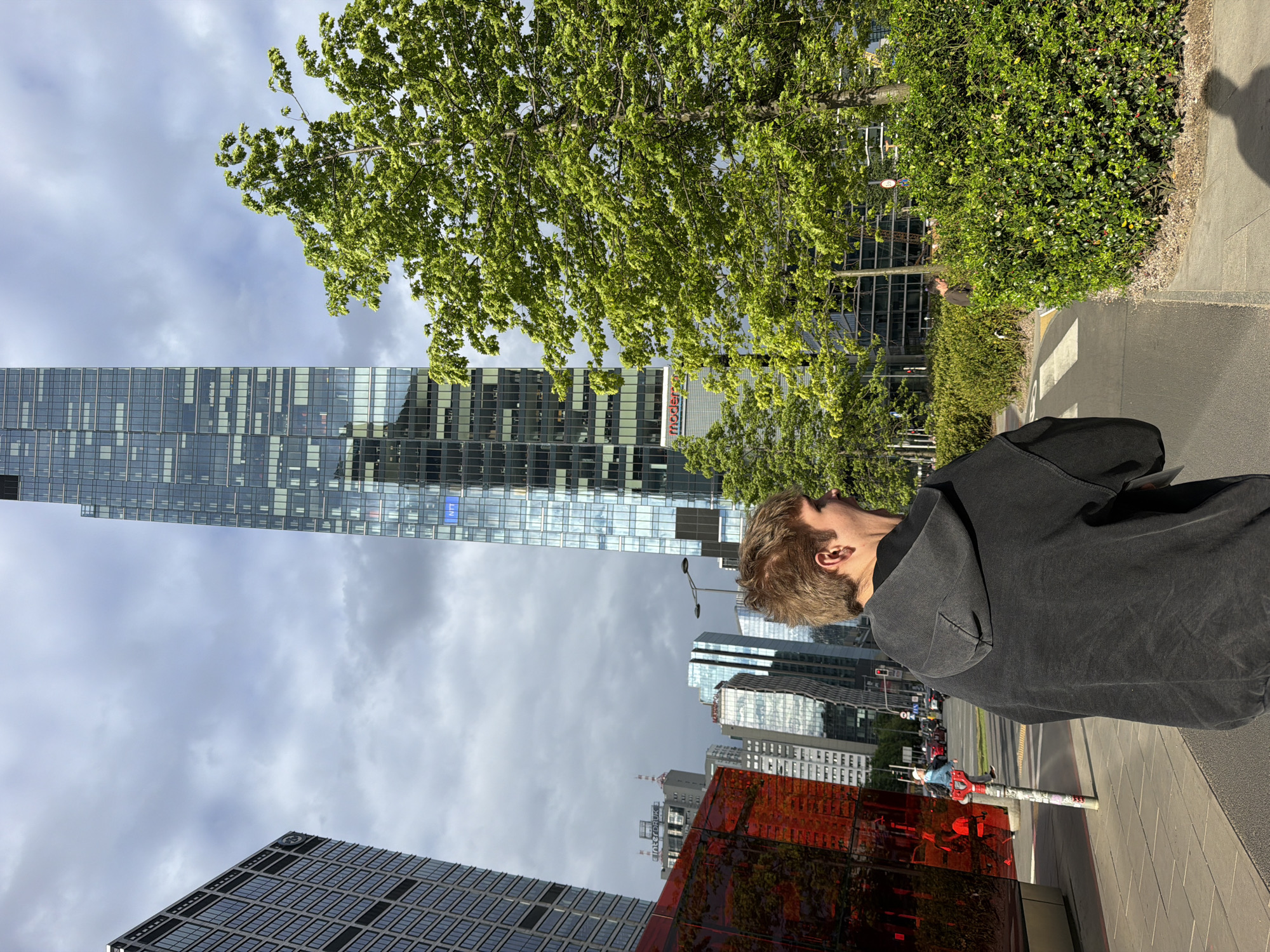 The image captures a person in a black hooded sweatshirt standing on a sidewalk in a city. The person's face is partially visible, and they are looking upwards. The background includes tall buildings with glass facades, one of which is notably orange. Trees and greenery are interspersed among the buildings. The sky is cloudy.