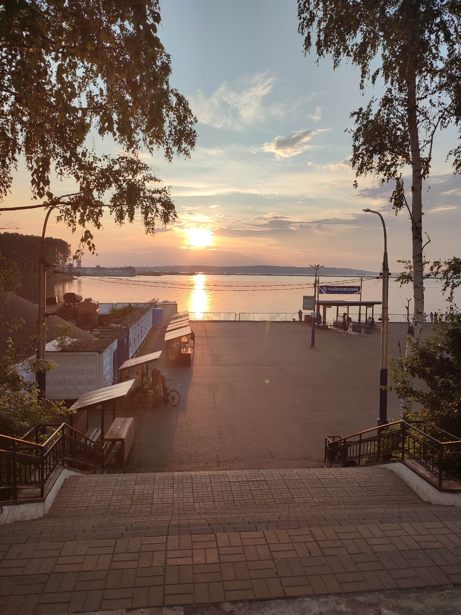 The image depicts a waterside promenade at sunset. The sun is setting over a wide body of water, casting a warm glow and a reflection on the surface. In the foreground, a set of brick stairs leads down to the promenade. The promenade itself is paved and features several covered stalls or kiosks, some with benches. Two bicycles are parked near one of the stalls where two people are seated. Further along the promenade, there is a bus stop shelter with a sign that reads "ЧАЙКОВСКИЙ" (Tchaikovsky). A lamppost stands near the bus stop. On the left side of the image, there are some large containers and piles of gravel. Trees with branches extending into the frame are visible on both the left and right sides. In the distance, across the water, a silhouetted shoreline with trees and some industrial structures can be seen, along with a ship on the water. The sky is filled with soft clouds and the warm hues of sunset. The overall atmosphere is tranquil and serene.