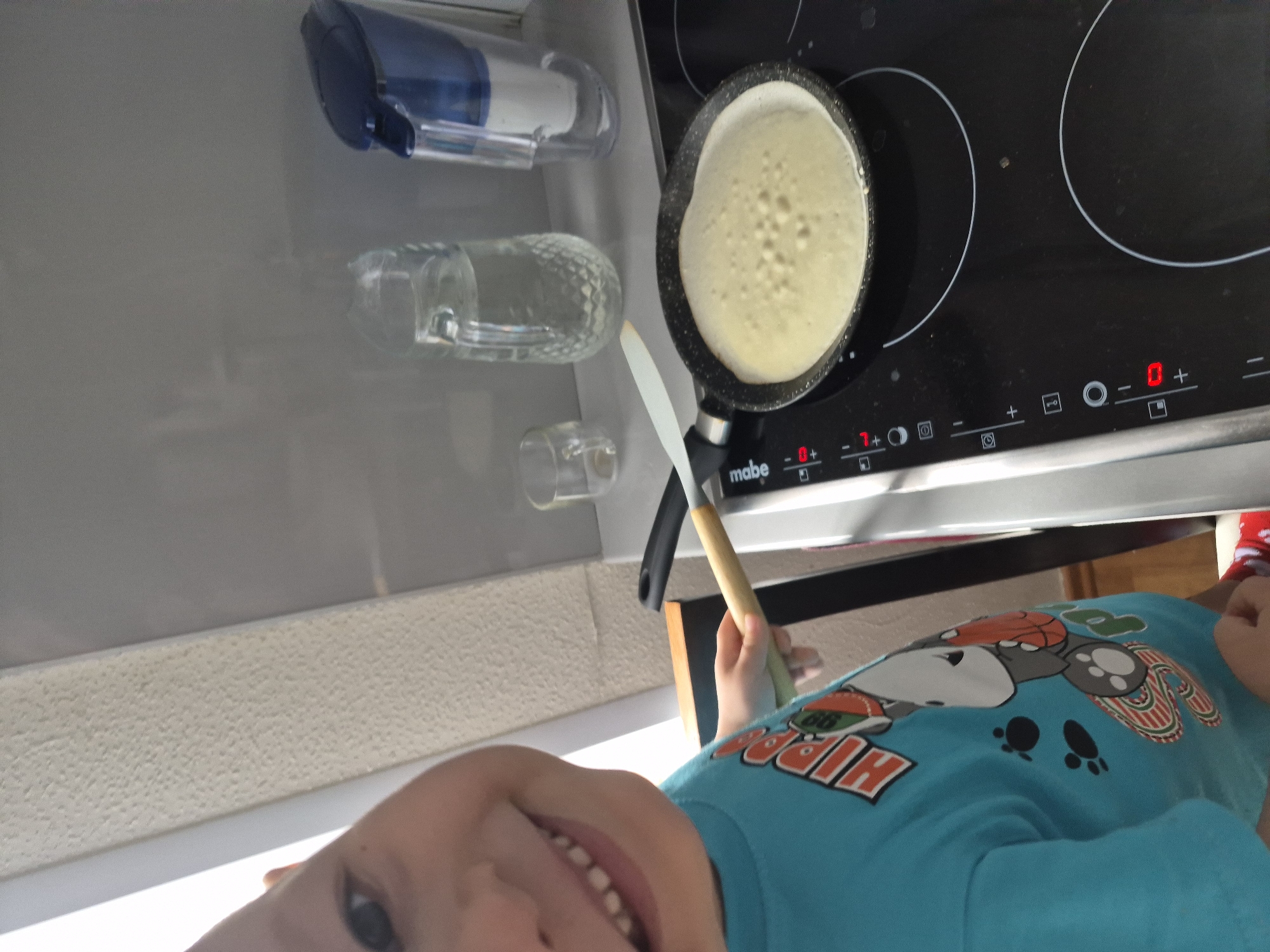 A child is making a pancake in a kitchen. The child, wearing a blue shirt, is holding a spatula and smiling. A pancake is cooking in a black frying pan on a black stovetop. The stove is marked with "mabe". A blue and white container and two glass containers are on a counter next to the stove. The setting is a kitchen but further details of Tobolsk, Russia are not discernible from this image.