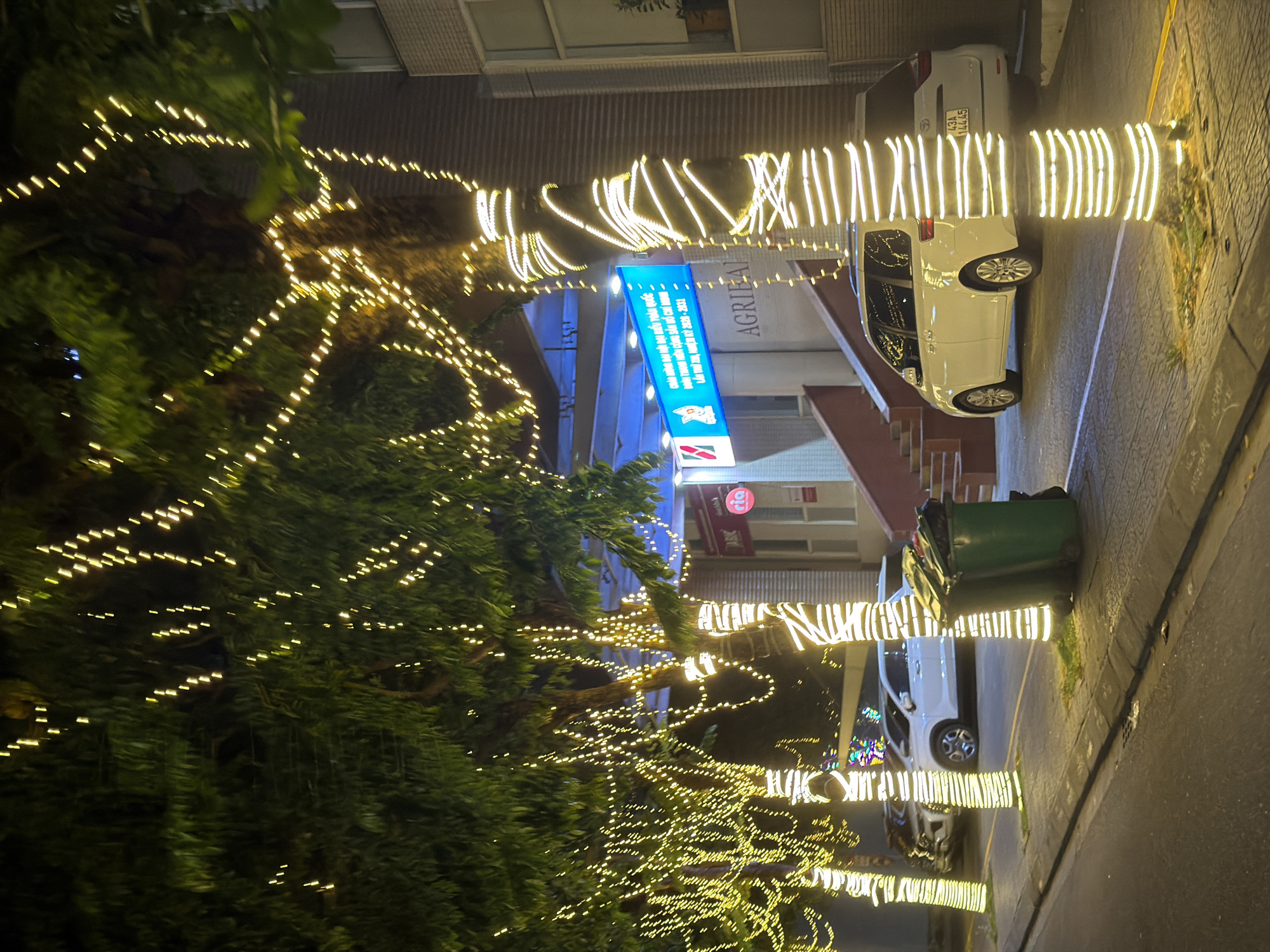 The image shows a street scene at night in Da Nang, Vietnam, likely near the Dragon Bridge due to the festive lighting. Trees lining the sidewalk are elaborately decorated with strings of yellow lights, creating a festive and warm ambiance. A white minivan is parked on the street, and in the background, a building with signage that appears to be in Vietnamese, possibly advertising a business or event, is visible. The street is paved, and there are two green trash bins on the sidewalk. The lighting suggests it could be a special occasion, like a holiday or festival.