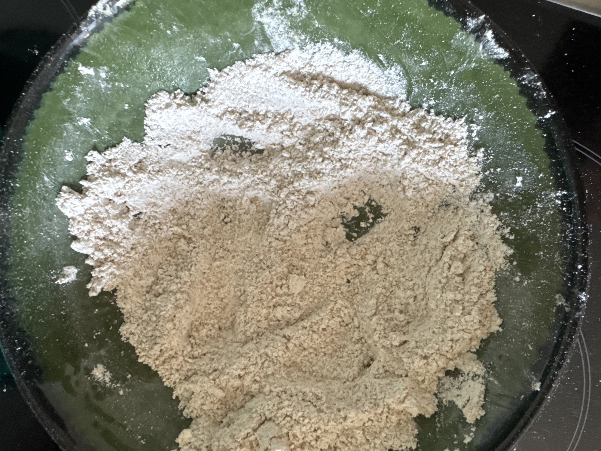 The image shows a pile of light brown powder, possibly flour or a similar dry ingredient, spread across a green bowl. The powder is unevenly distributed, with some areas thicker than others and a scattering of fine particles on the green surface of the bowl. The bowl itself appears to be ceramic with a textured or slightly glossy finish. The background is dark, suggesting it might be a countertop or a stovetop. The scene is indoors, and the lighting suggests it is daytime. There are no people or animals visible in the image. The image is cropped closely to the bowl, with no wider context of the surroundings. There is no information available about the location within Höhenkirchen-Siegertsbrunn, Germany.