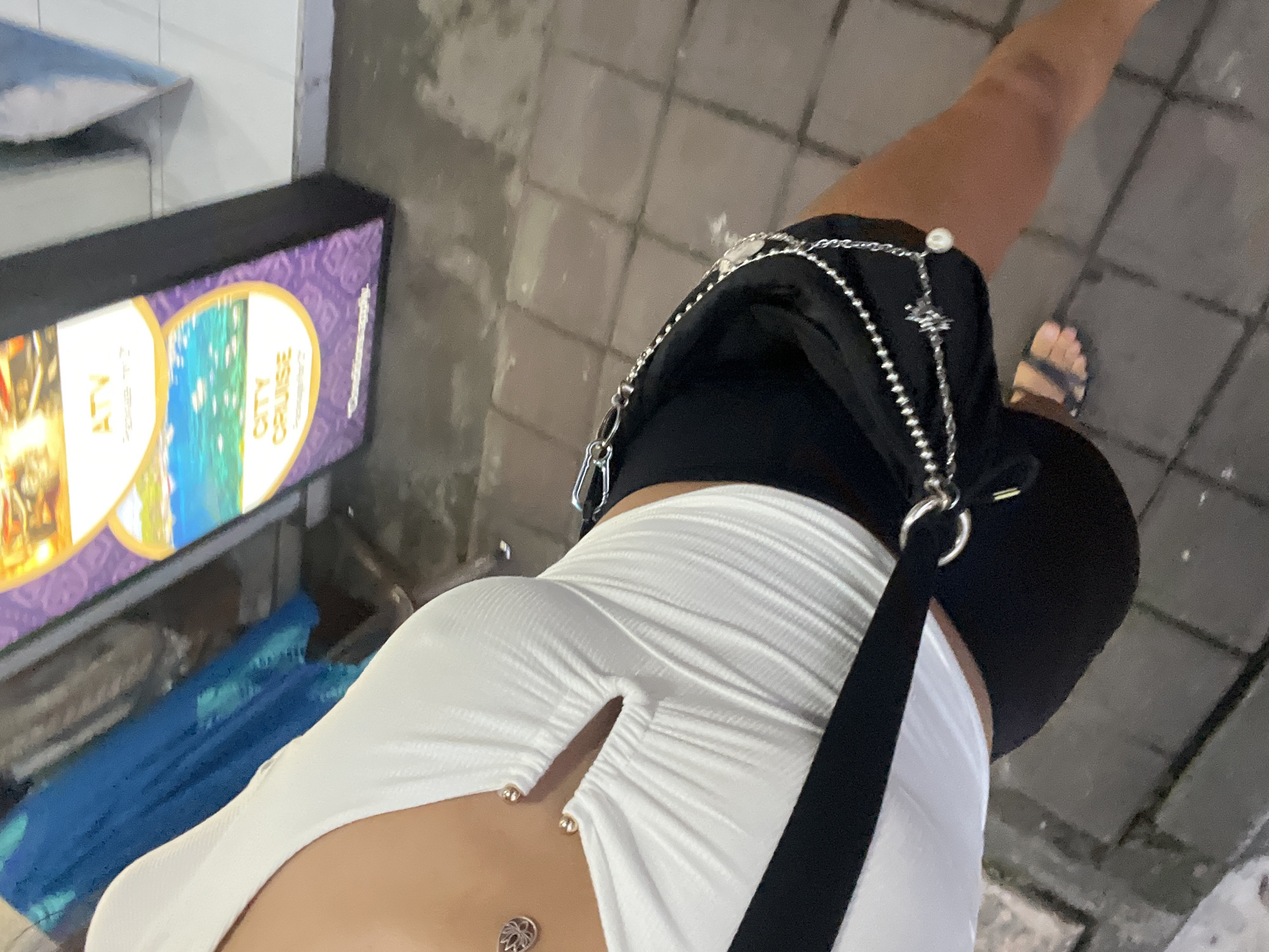 A high-angle shot shows a person on a paved street in Kuta Selatan, Indonesia. The person is wearing a short, white, ribbed dress with a slit at the front and a belly button piercing visible. The person also wears black shorts and sandals on their right foot. A black purse with a silver chain strap is slung over their shoulder. To the left of the person is a sign advertising "ATV" and "City Cruise." A blue object is visible beneath the sign. The background is the street.