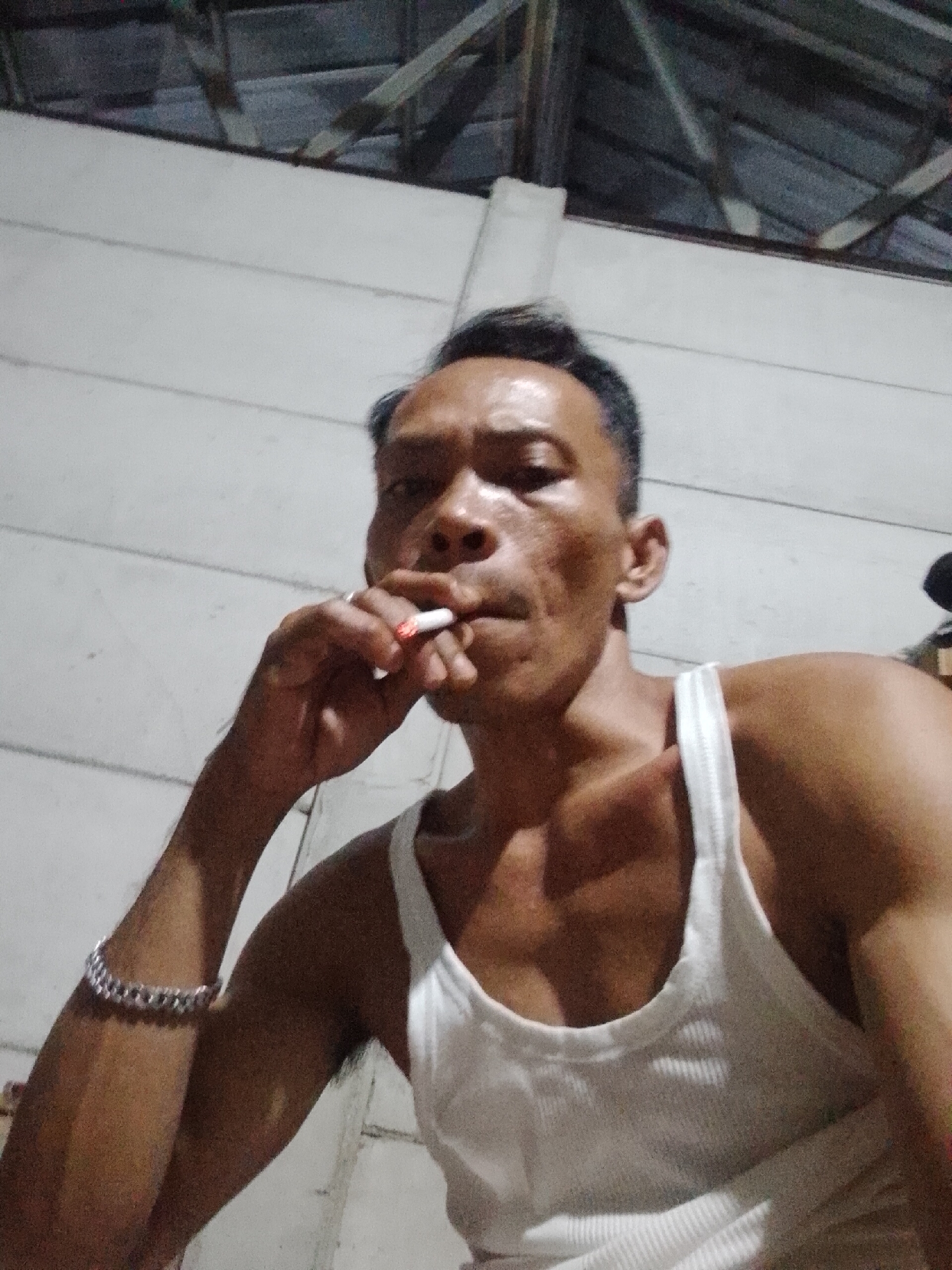 A medium shot depicts a man in a white tank top smoking a cigarette indoors. The background consists of a white wall and a partial view of a ceiling with wooden beams. The man wears a silver bracelet. The location is Unknown, Unknown.
