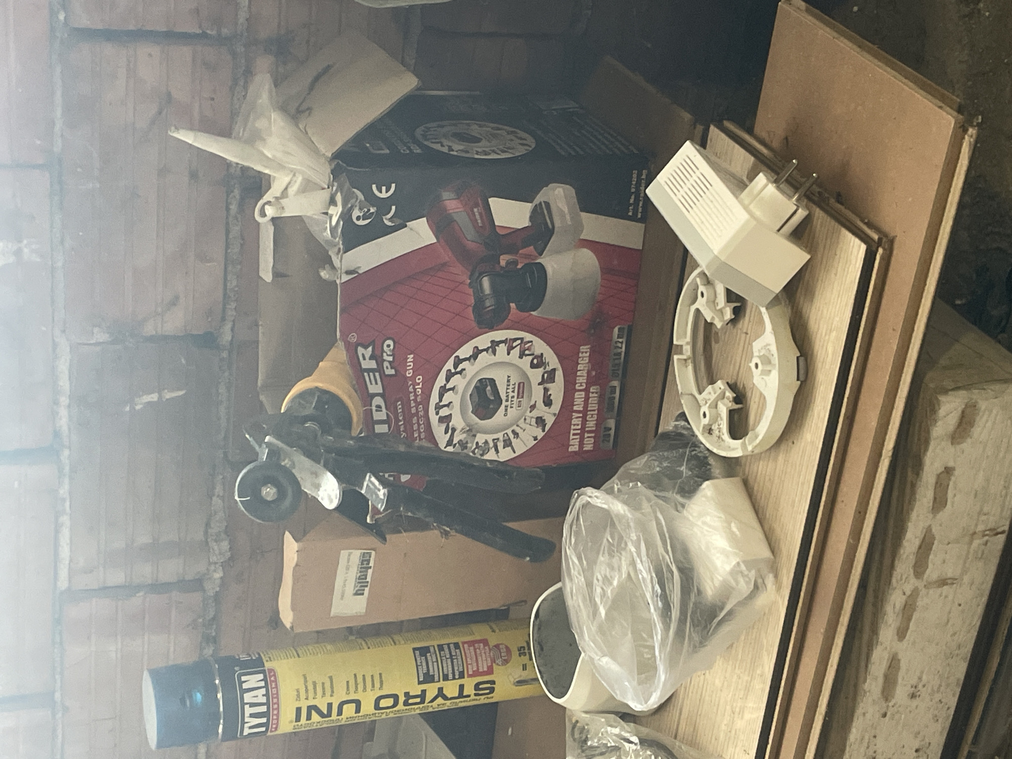 The image captures a cluttered storage area, likely a workshop or garage, illuminated by a bright light source from the left, causing glare. The scene is dominated by various objects, tools, and construction materials.
In the center, a red and black cardboard box for a "RAIDER Pro CORDLESS SPRAY GUN SGC20 SOLO" is prominent. The box states "ONE BATTERY FITS ALL" and "BATTERY AND CHARGER NOT INCLUDED," indicating it's for a 20V tool with 1000ml capacity and nozzle sizes D1.5, 18, and 22mm. It also displays "Art. No. 074202," "www.raider.bg," and a "CE" mark.
To the left of the box, a yellow and blue can of "TYTAN PROFESSIONAL STYRO UNI" spray foam is visible, with partially legible Bulgarian text including "Ziduri" (Walls), "Acoperisuri" (Roofs), and "=35." Above it, a yellow and black tool, possibly an angle grinder, is partially seen, along with a smaller brown box that might read "Scholly."
To the right of the central box, several items are placed on wooden boards. These include a white electrical adapter/plug with two prongs, a white circular plastic base (possibly for a smoke detector or similar device), and a clear plastic bag containing an unidentified white object.
The background shows a rough, possibly brick, wall. The overall impression is one of utility and disarray, typical of a storage space for work-related items. No human figures are present, and there is no discernible activity taking place. The time of day is not indicated due to the indoor setting and lighting conditions. Given the location context, the Bulgarian text on the TYTAN can is consistent.