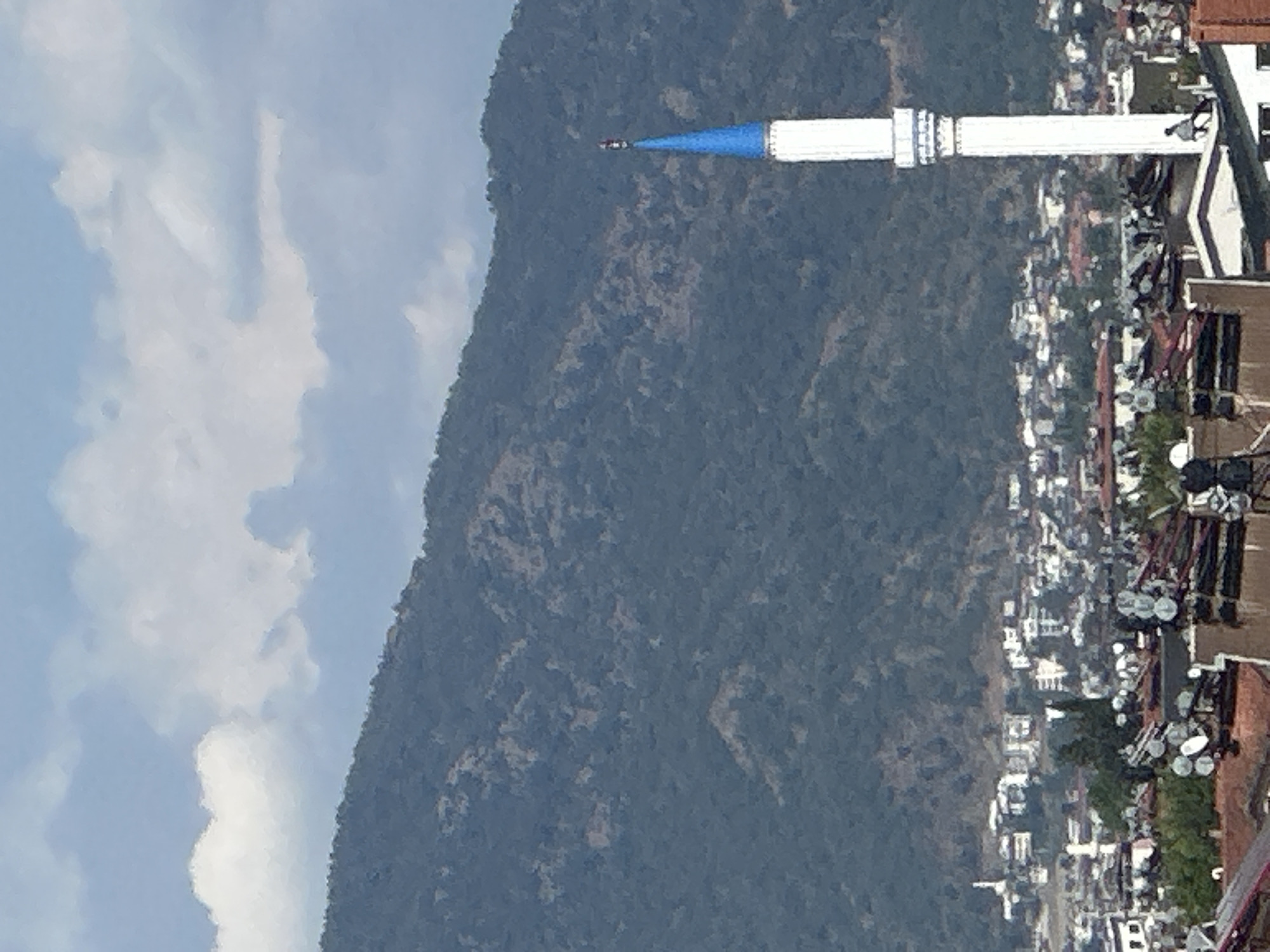 The image shows a scene in Fethiye, Turkey. In the foreground is a cluster of buildings, possibly residences, with many satellite dishes visible. Extending from the right side of the frame is a slender, tall, white minaret with a blue cone-shaped top. Behind the minaret is a densely forested mountain. To the left of the mountain are clouds in a blue sky. The image appears to be taken from an elevated position, looking down onto the town and the minaret, with the mountain as a backdrop.