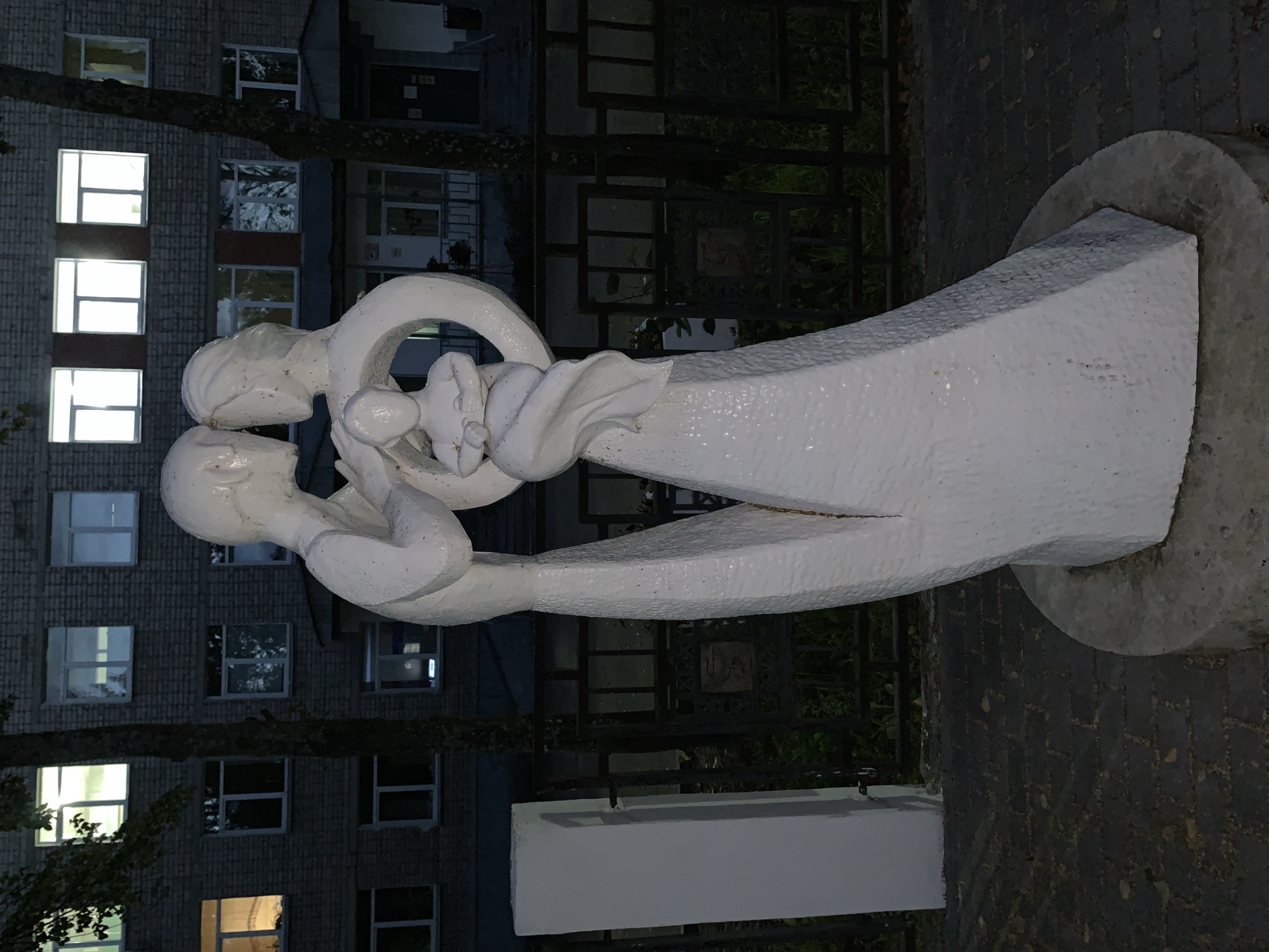 This is a medium shot, eye-level, outdoor photograph of a white sculpture depicting a family with a child. The sculpture is located in front of a brick building with multiple windows. The building is illuminated from within, casting a soft glow. A dark metal fence runs horizontally behind the sculpture. The ground in front of the sculpture is paved with dark interlocking bricks, and a white rectangular pillar stands to the left. The sculpture itself is abstract, with smooth, flowing lines. The figures are simplified, with the parents embracing and holding a child between them. The child is depicted as a smaller, rounded form. The sculpture is situated on a circular concrete base. The overall atmosphere of the image is serene and contemplative, likely taken at dusk or dawn due to the ambient light. The location is identified as Chaikovsky, Russia.
