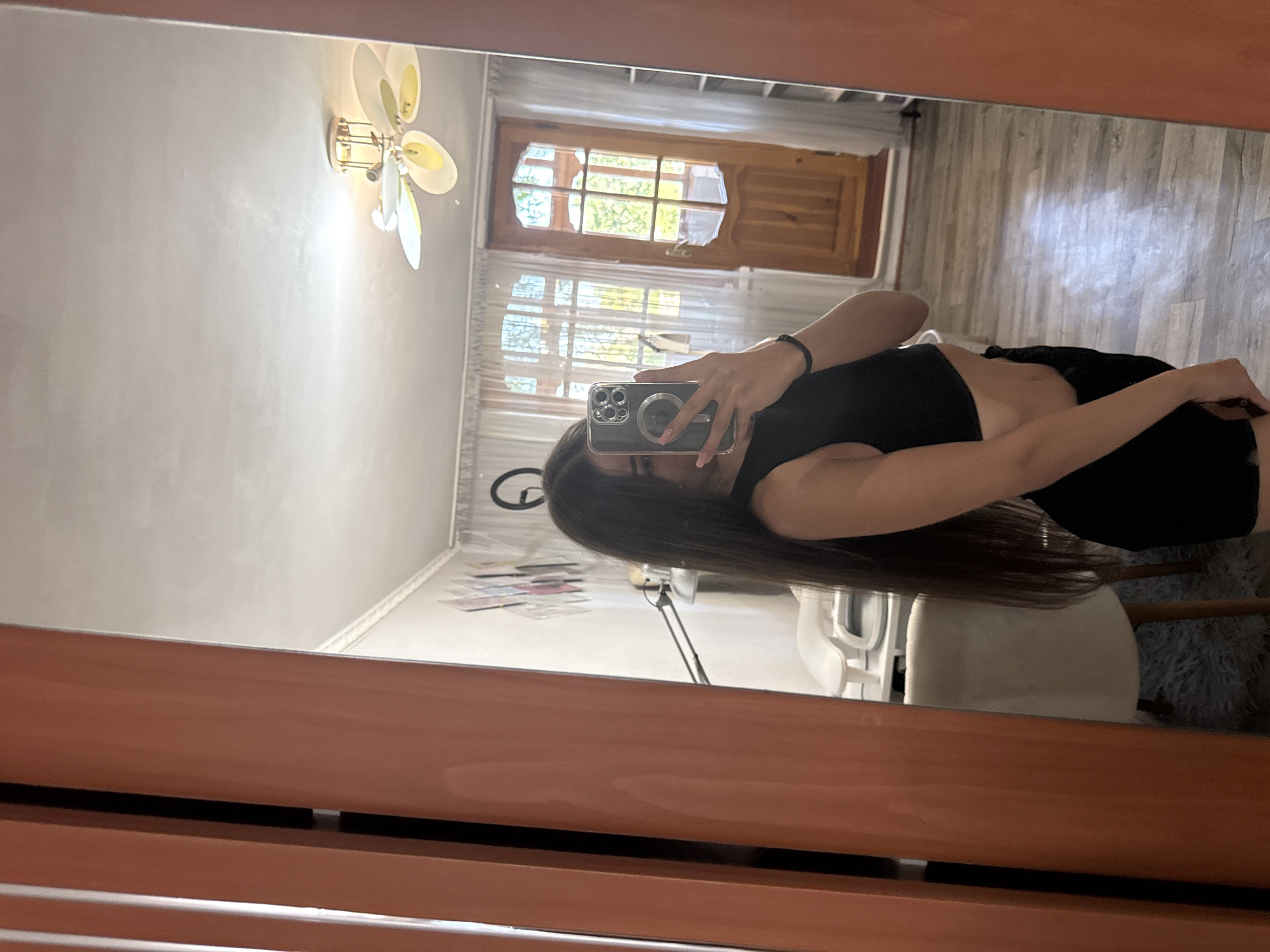 A young woman with long, dark hair takes a mirror selfie in an indoor setting. She is wearing a black crop top and black shorts. She holds a smartphone in her right hand, capturing her reflection. Behind her, a room is visible with a ceiling fan, a wooden door, and windows with white curtains. A chair with a light-colored cushion is also present. The overall impression is of a casual, personal moment captured within a home. The image does not provide specific visual clues to pinpoint a location within the city of Basarabeasca, Moldova.