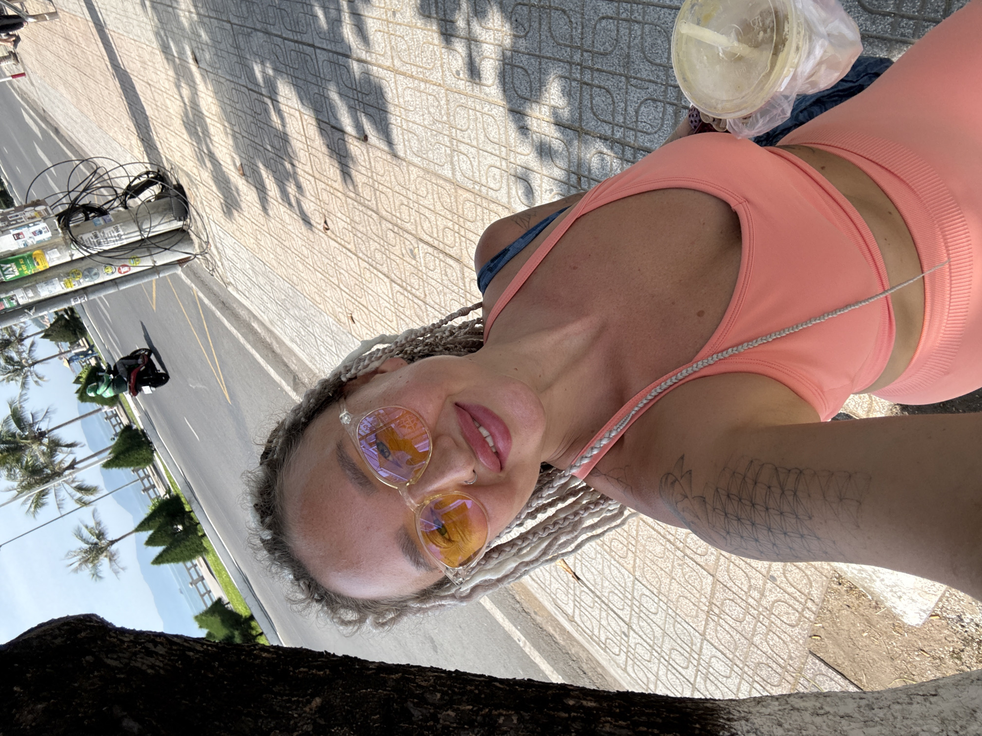 A woman with blonde braided hair and orange-tinted sunglasses smiles at the camera, holding a drink. She is wearing a coral-colored tank top and matching leggings. Her left arm, visible in the foreground, has a black line-art tattoo. The woman stands on a sidewalk paved with square tiles, with a tree trunk on the left side of the frame. Behind her, a road with a few cars and palm trees can be seen. The location is identified as Bac Nha Trang Ward, Vietnam.