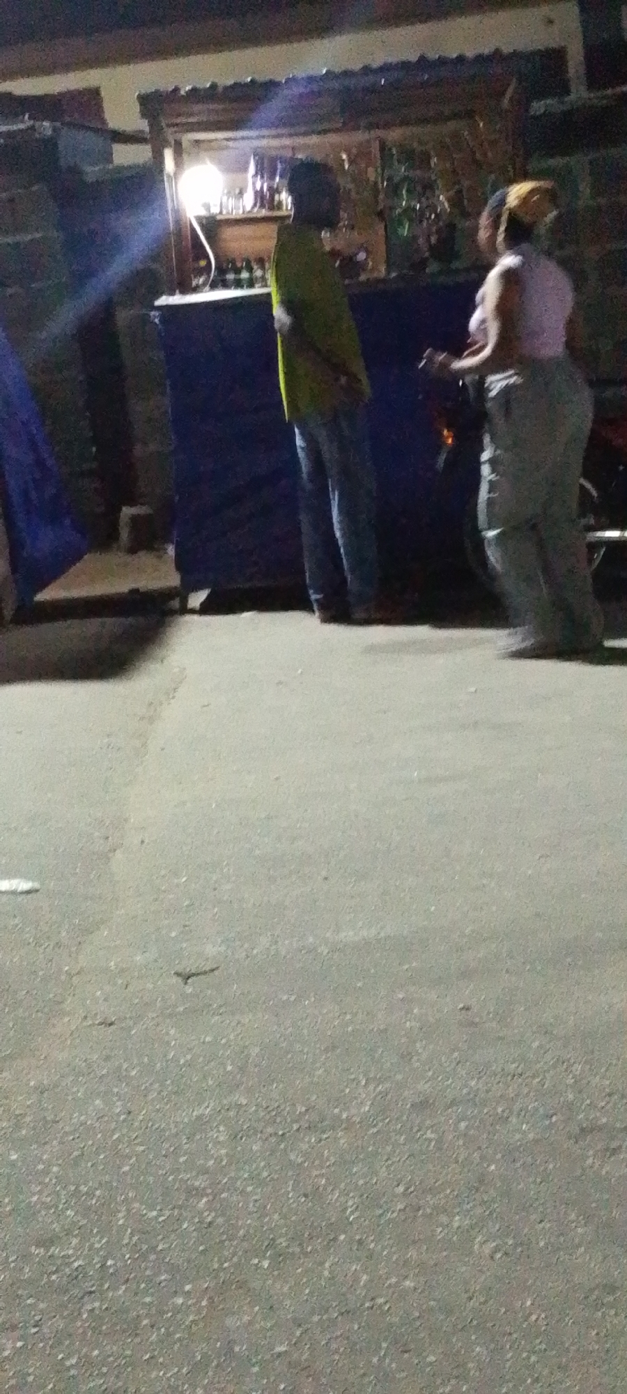 The image captures a street scene in Lokoja, Nigeria, at night. A makeshift bar stands to the right of the frame, illuminated by a light bulb. A man in a yellow shirt and jeans stands near the bar. A woman wearing a light-colored top and pants is next to him. They are talking or interacting in some way. A motorcycle is parked near the bar. To the left, a blue tarp is visible. The ground is paved.