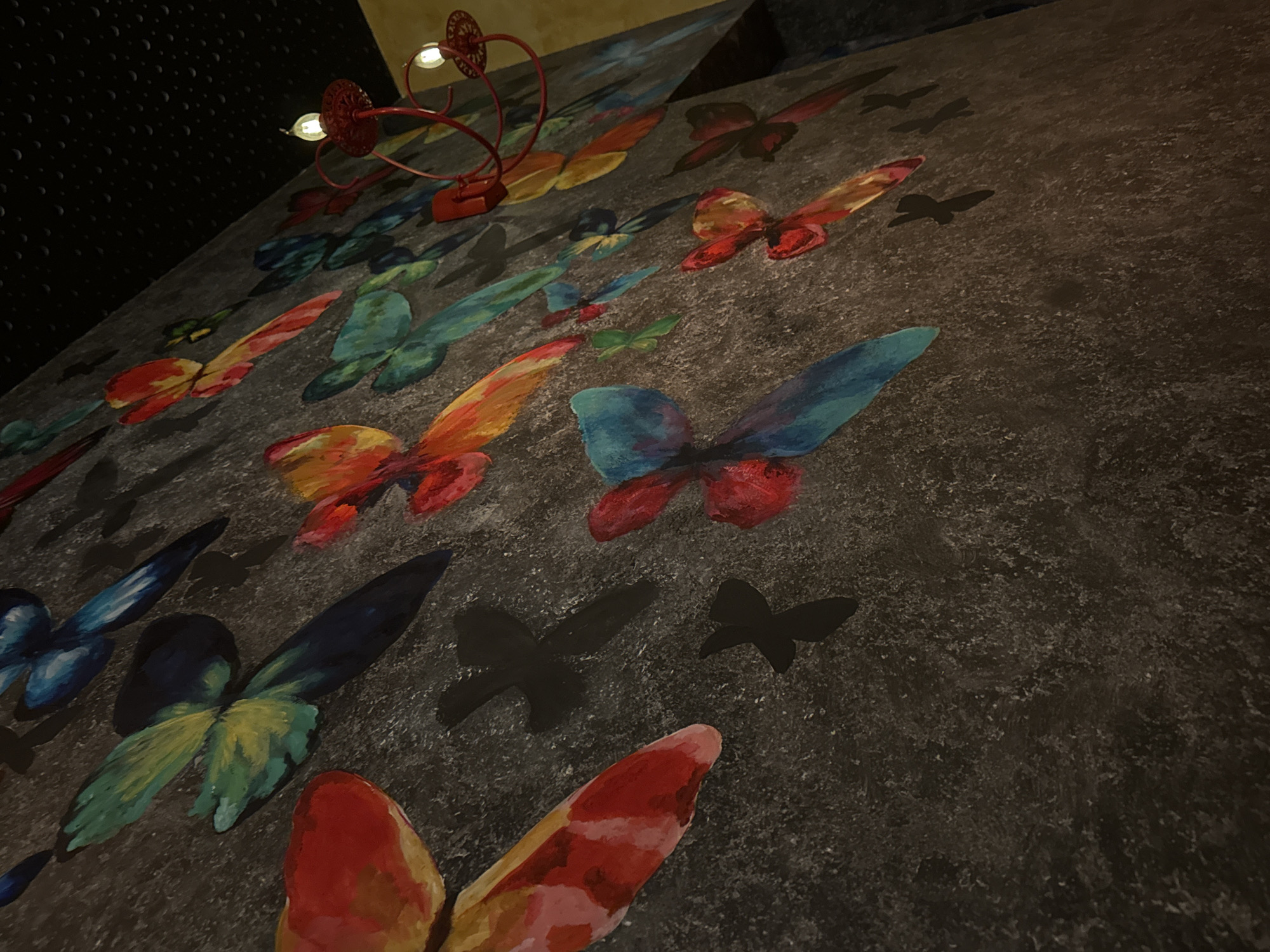 The image displays an indoor scene focused on a decorative wall. The wall surface is dark grey with a coarse, mottled texture, resembling painted concrete or stucco. Adorning the wall are numerous painted butterflies in various sizes and vibrant colors including red, orange, yellow, teal, green, and blue. These butterflies are rendered in an abstract, fluid style with visible brushstrokes, some appearing in pairs or small clusters. Interspersed among the colorful butterflies are smaller, solid black silhouette shapes resembling flying birds or abstract butterfly forms, painted directly onto the wall.
On the right side of the image, a red, ornate, wrought-iron style lighting fixture is visible, extending from the wall or ceiling. Two small, clear, incandescent light bulbs are illuminated on this fixture. Above the light fixture, a portion of the ceiling is visible; it is dark and features a textured, possibly dimpled or perforated pattern. No individuals are present in the scene, and no specific actions are depicted. The location in the city of Sofia, Bulgaria, cannot be determined from the visual content of the image.