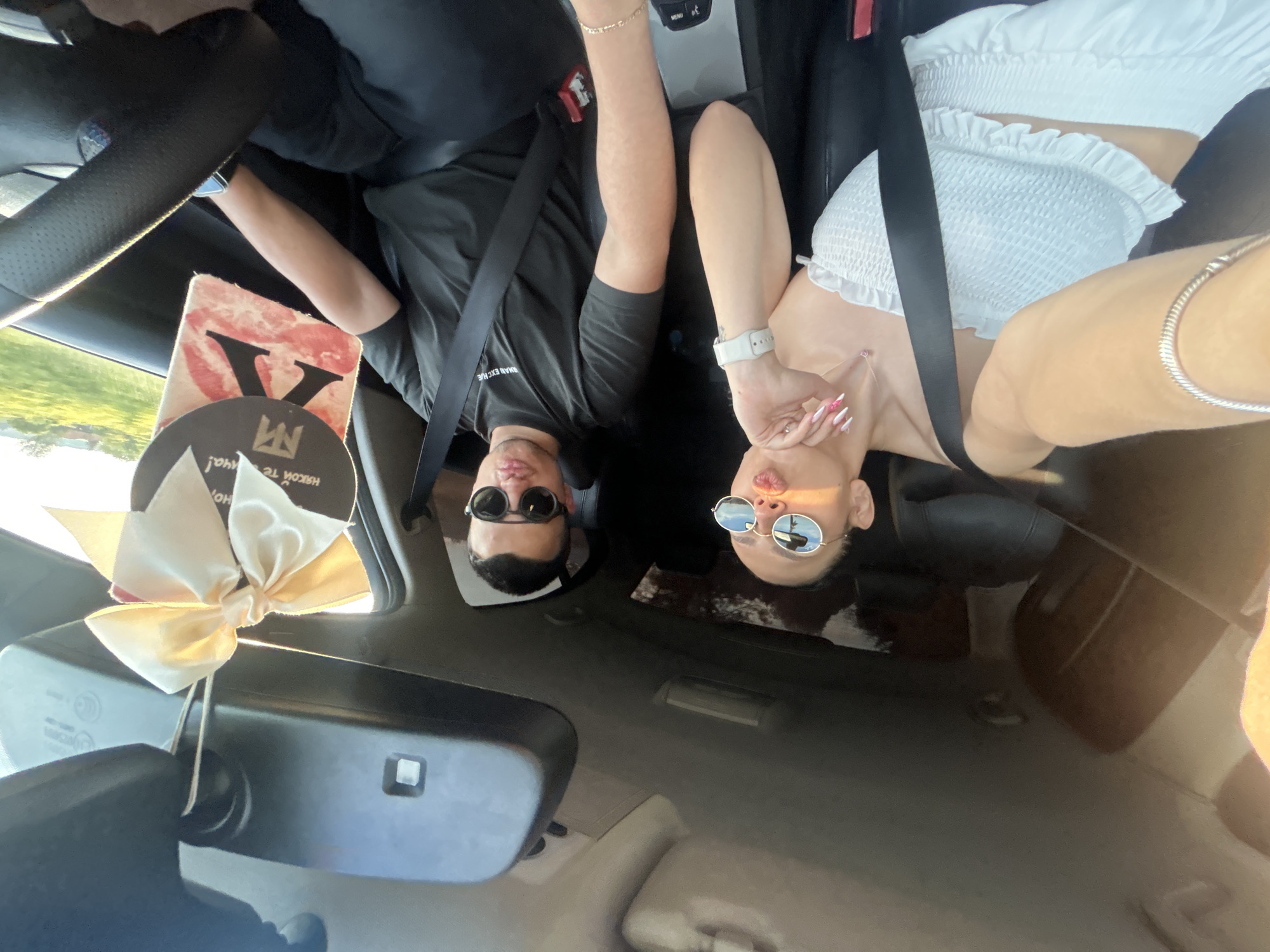 A selfie taken inside a car, angled to show the front seats, two people, and parts of the dashboard and windshield.
Two individuals, a male and a female, are visible in the front seats. Both are wearing sunglasses and seatbelts. The male is wearing a black shirt, while the female is wearing a white top.
On the dashboard, partially visible, is a gift box or package with a ribbon, and a paper card with a round coaster with logos.
Through the windshield, there is a glimpse of what appears to be a road or open field, indicating an outdoor environment.
There is no specific information in the image to pinpoint a location within the city of Borovo, Bulgaria.