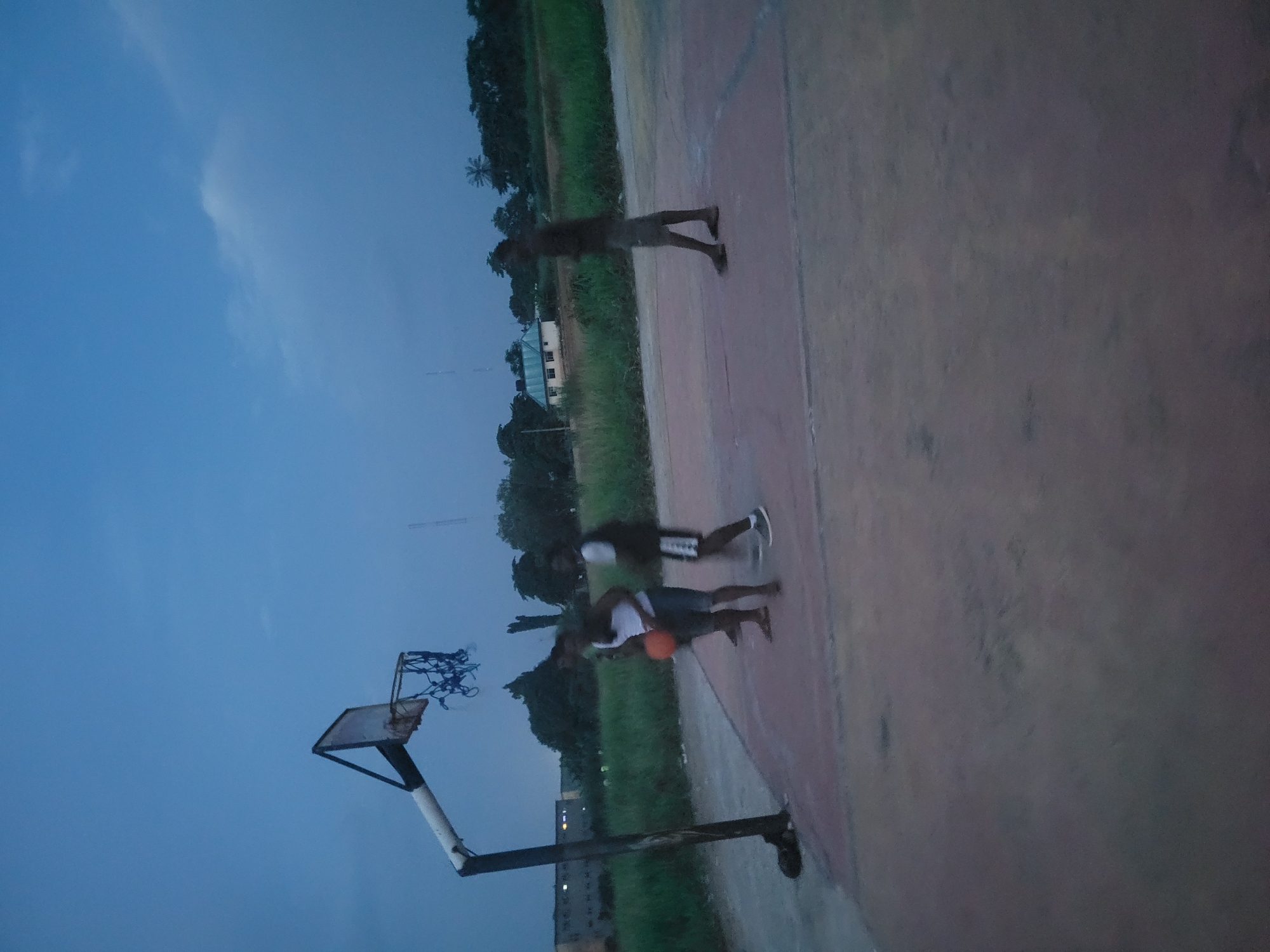 The image shows a basketball court in Egbagu, Nigeria, with a group of people playing basketball. The scene is set outdoors, likely in the late afternoon or early evening as indicated by the dim lighting and the blue sky with some clouds. The basketball court surface appears to be made of concrete or asphalt, and there is a basketball hoop with a damaged net.
There are at least three individuals visible in the image, all of whom appear to be young men. They are dressed in casual athletic wear. One person is in the process of dribbling or passing a basketball, which is orange. Another individual is standing nearby, seemingly observing or participating in the game. A third person is further away, in the background, standing near the edge of the court, possibly waiting to join in.
The surrounding area features lush green vegetation, including trees, suggesting a tropical or subtropical climate. In the background, some buildings and what appears to be a radio tower or a similar tall structure are visible, offering clues about the urban or semi-urban setting. The overall mood seems to be one of casual recreation and sport. The damaged net on the basketball hoop is a notable detail, indicating that the court might be well-used or subject to wear and tear. No text is visible in the image.