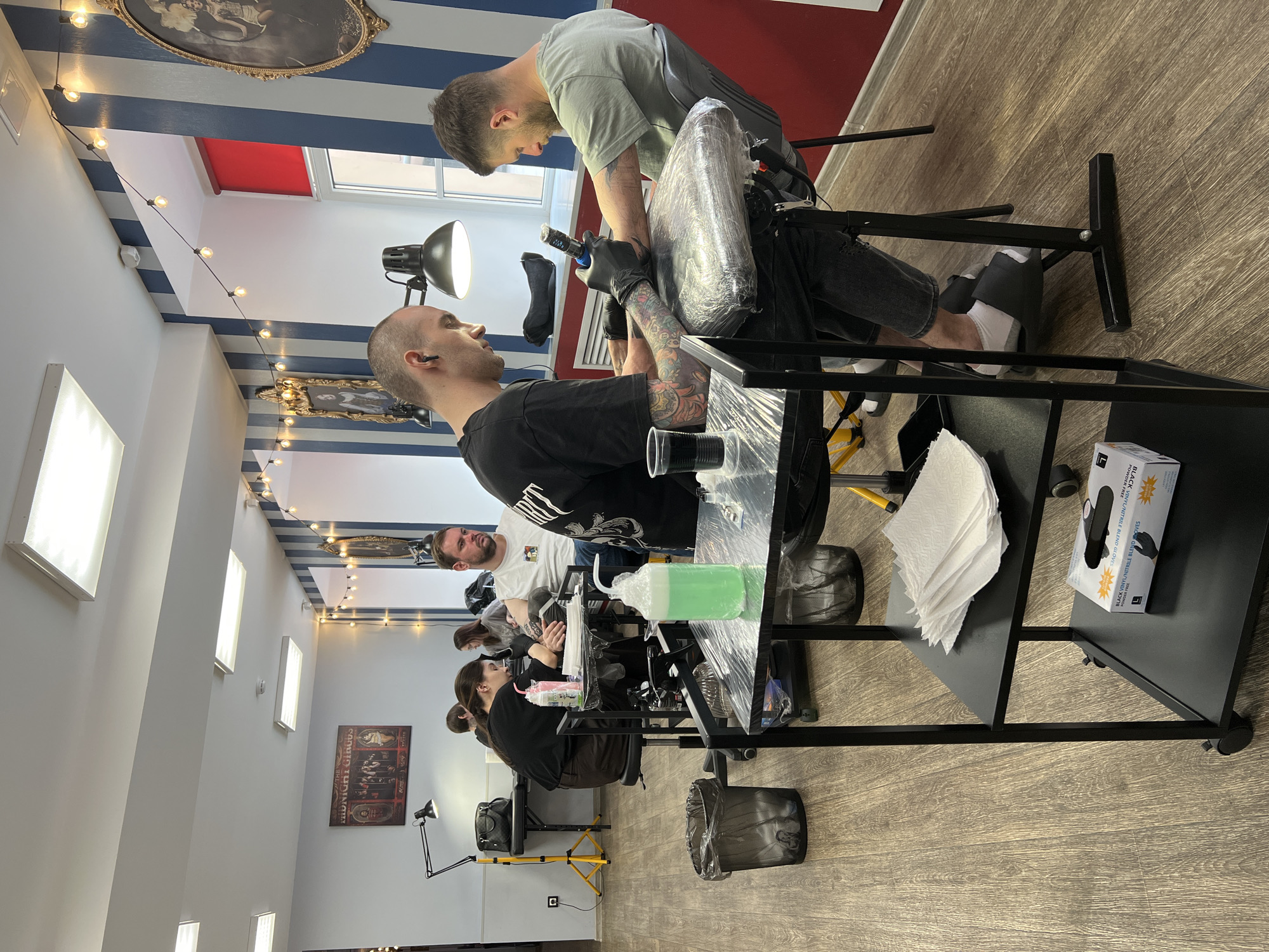 The image captures an active tattoo studio in Saint Petersburg, Russia, featuring several individuals engaged in tattooing. In the foreground, a tattoo artist, wearing a light grey t-shirt and black gloves, leans over to tattoo the bicep of a client. The client, dressed in a black t-shirt, sits in an adjustable chair, looking towards the artist. His arm, heavily tattooed, rests on a padded support wrapped in plastic for hygiene.
The artist's immediate workspace includes a black trolley table holding a bottle of green liquid (likely disinfectant or soap), paper towels, and a box of black vinyl/nitrile blend gloves. The table is also covered in plastic wrap. A task lamp illuminates the work area. The client receiving the tattoo has visible tattoos on both arms and an earpiece in his right ear.
In the background, several other tattoo artists and clients are visible, seated at similar workstations. One man in a white t-shirt and dark beard is attending to a client further back. The studio boasts a distinctive decor with a ceiling painted in blue and white stripes, adorned with string lights. A large window allows natural light into the space, with a red element visible beyond it. Framed artwork hangs on the walls, including a poster that appears to read "THE MIDNIGHT CIRCUS" in the mid-ground. The floor is made of light-colored wood laminate. The overall atmosphere is busy and professional, with a focus on precision and hygiene.