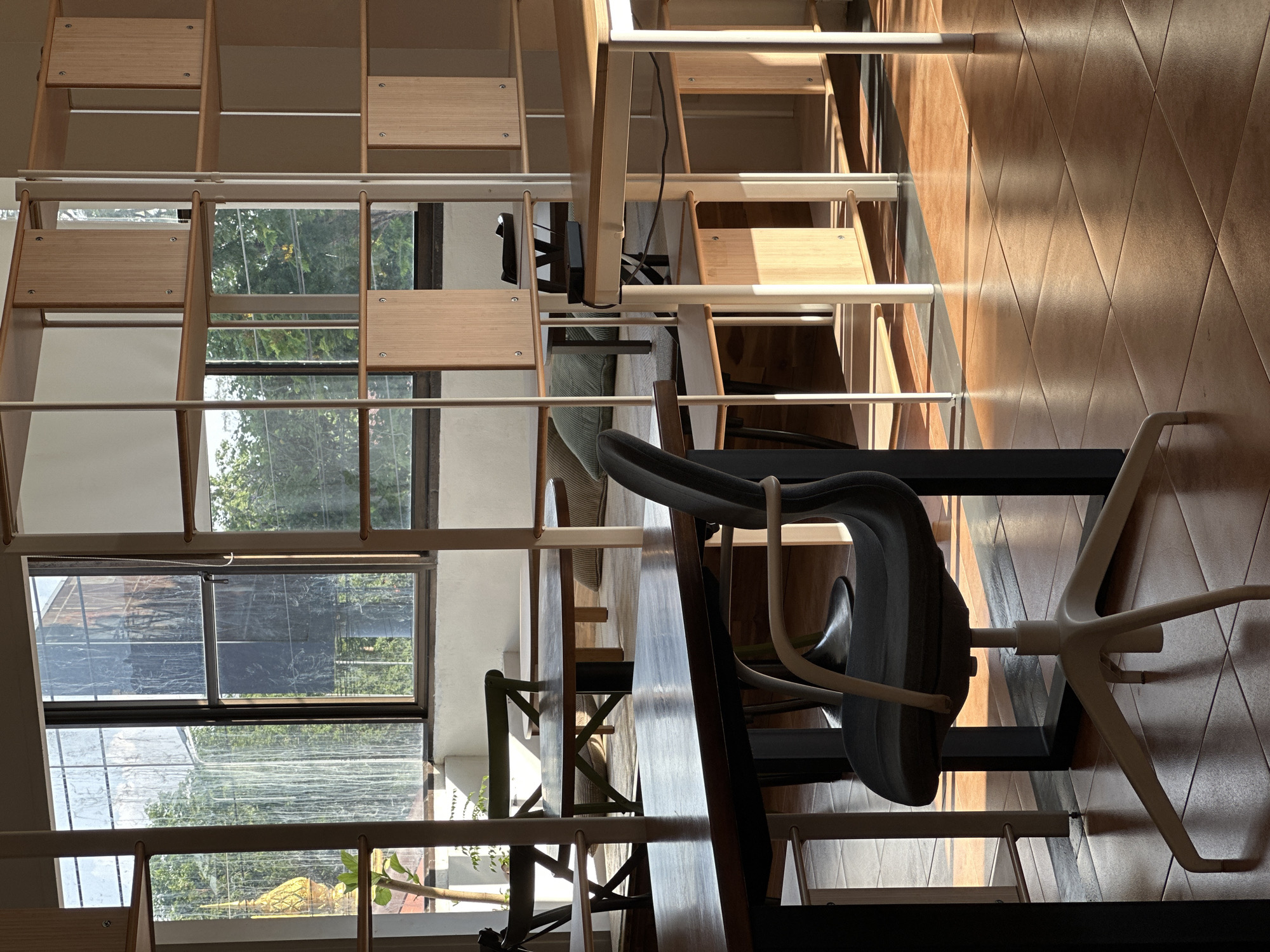 The image shows an indoor cafe or co-working space with large windows that let in natural light. The sun casts long shadows across the tiled floor. There are several tables and chairs, some of which are arranged in a seating area with cushions. A modern shelving unit with wooden shelves stands against a wall. Through the windows, trees and a golden spire can be seen, suggesting the location is in a tropical region. The overall atmosphere is bright and airy, with a focus on natural light and wooden elements. There are no people visible in the image. The specific location in ตำบลเกาะพะงัน, Thailand is an interior space, likely a cafe or coworking area, characterized by its open design, abundant natural light, and warm, earthy tones from the wooden furniture and tiled flooring. The view through the windows suggests proximity to lush greenery and possibly a religious structure, indicative of the local environment.