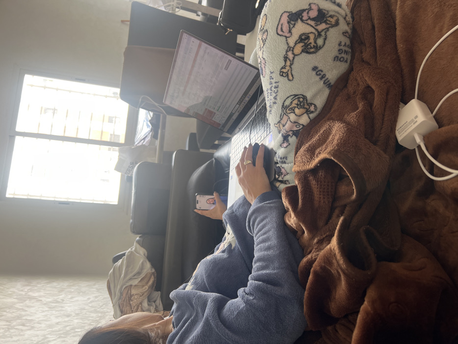 In Skhirat, Morocco, an indoor scene depicts an individual seated, partially obscured from the rear and side, engaged with electronic devices. The person, appearing to be female, wears a blue fleece top and has dark hair. A ring is visible on the fourth finger of her left hand. She operates a silver-toned laptop with her right hand on an external mouse, while holding a smartphone in her left hand, which displays a female face, indicative of a video call or image. The laptop screen shows what appears to be a spreadsheet application.
The individual is covered by a brown, textured blanket, underneath which a light blue pillow adorned with cartoon characters, including Smurfette and a character with "#GRUMPY FACE!", is visible, supporting the laptop. A white power adapter with a cable rests on the brown blanket in the foreground.
The environment includes a dark brown or black leather sofa or armchair behind the person. A large window with white frames and horizontal bars dominates the background, admitting significant natural light, partially overexposing the exterior view, which suggests another building. A white shopping bag labeled "PRESS" is situated near the window. The wall to the left features a textured, light-colored finish. Additional furniture, including other dark chairs, is partially visible in the background.