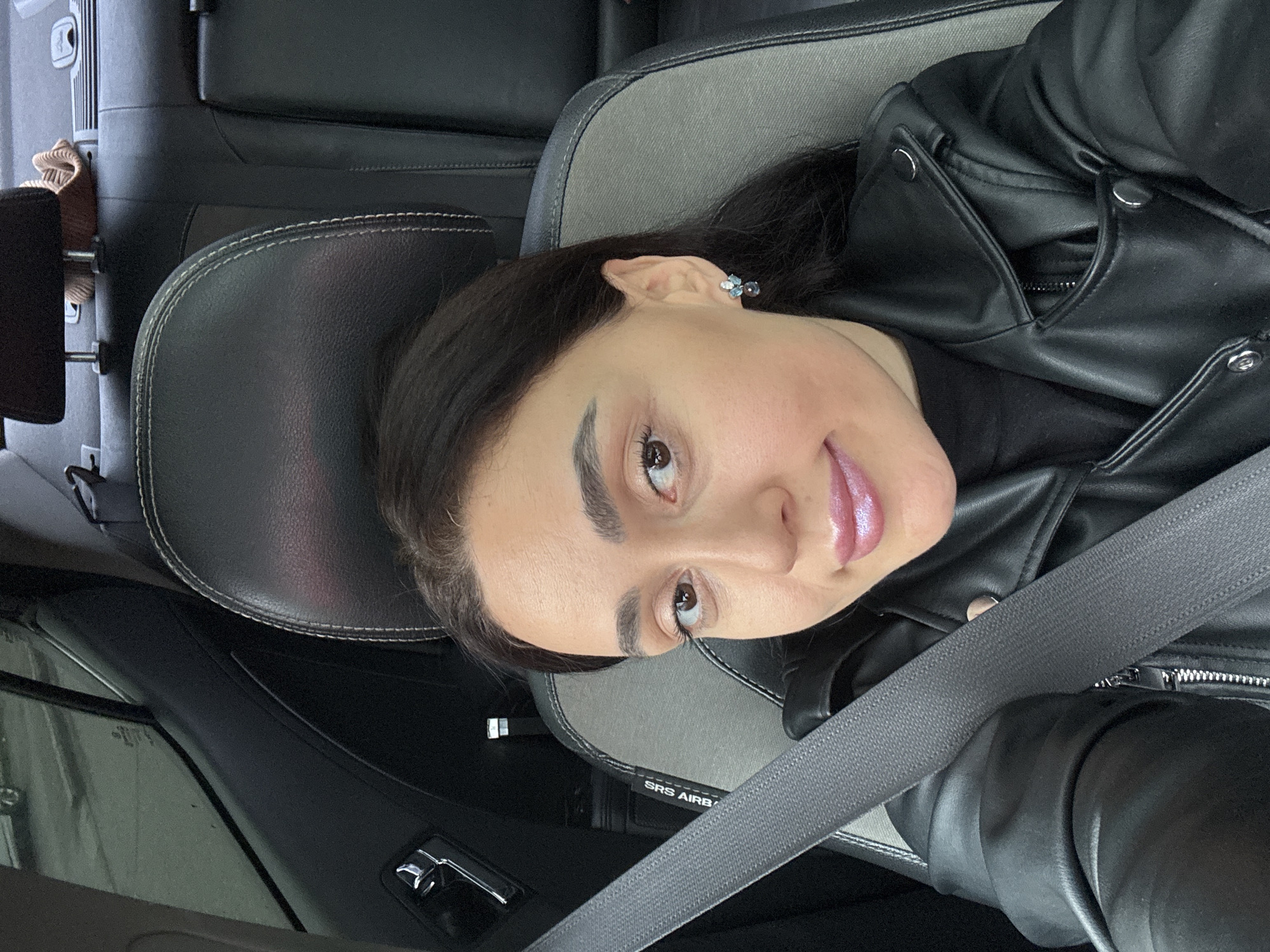 An adult female is depicted in a close-up selfie taken from within a vehicle. She has dark hair, brown eyes, filled eyebrows, and is wearing makeup, including shimmering pink lip gloss and winged eyeliner. She is smiling faintly and looking directly at the camera. She wears a black faux leather jacket over a dark, possibly black, top and a small, multi-gemstone earring in her visible left ear. A grey seatbelt, labeled "SRS AIRBAG," is correctly positioned across her chest and shoulder. She occupies what appears to be the front passenger seat, which has black and grey fabric upholstery with white stitching on its black leather headrest. The car's interior includes a chrome door handle on the left side of the frame and dark-tinted windows revealing a blurred exterior street scene with a light-colored vehicle. The black rear seats are visible, with a light brown, possibly woven, object resting on the rear parcel shelf. This scene is situated inside a passenger vehicle in the city of Tbilisi, Georgia.