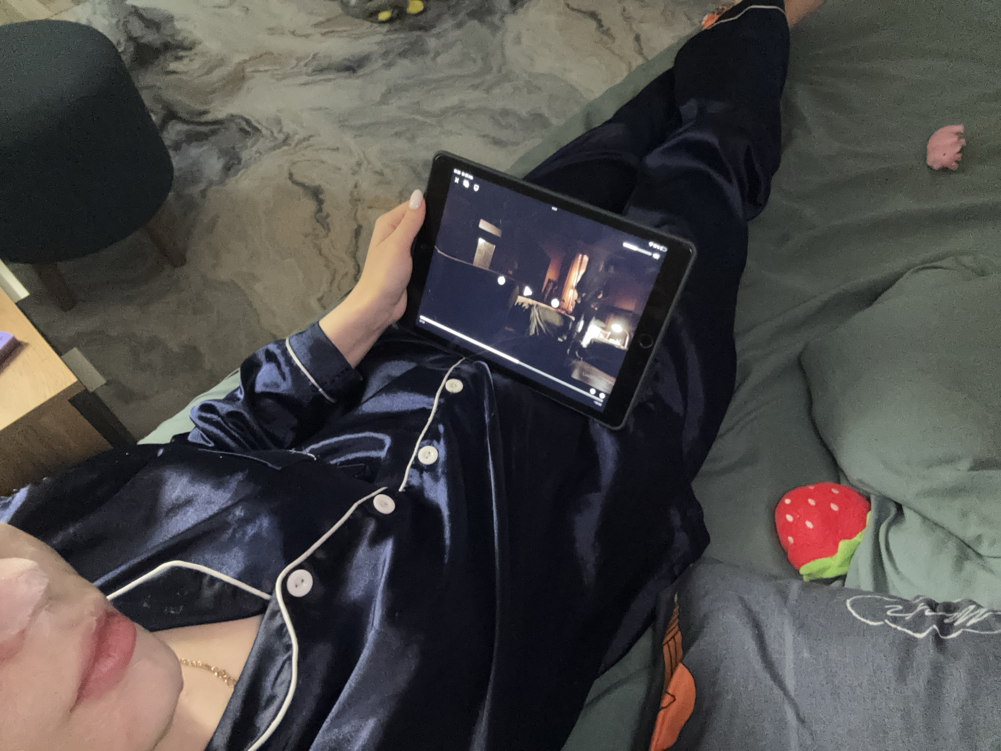 The image presents a top-down view of a person relaxing indoors, likely in Chaikovsky, Russia. The individual is lying down, dressed in comfortable, shiny navy blue satin pajamas featuring white piping and white buttons. A subtle golden chain is visible around their neck, and part of their face, possibly with a face mask applied, is seen in the lower-left foreground.
The person is holding a dark-colored tablet with their left hand, watching a video or movie. The content on the tablet screen displays a dimly lit indoor scene, possibly from a film, along with typical video playback controls such as a progress bar, play/pause icon, and status bar indicators for Wi-Fi and battery.
The setting appears to be a cozy home environment, possibly a bedroom or living room. The person is resting on a bed or couch covered with a combination of plain green and patterned grey bedding. On the green bedding to the right, a small pink figurine, possibly a pig, is visible. Further down, nestled in the grey bedding, is a red and green strawberry-shaped plush toy. In the background on the left, a dark chair or stool with wooden legs sits on a multi-toned, patterned rug or carpet. The overall lighting is subdued, suggesting it is either evening or nighttime, contributing to a relaxed and comfortable ambiance.