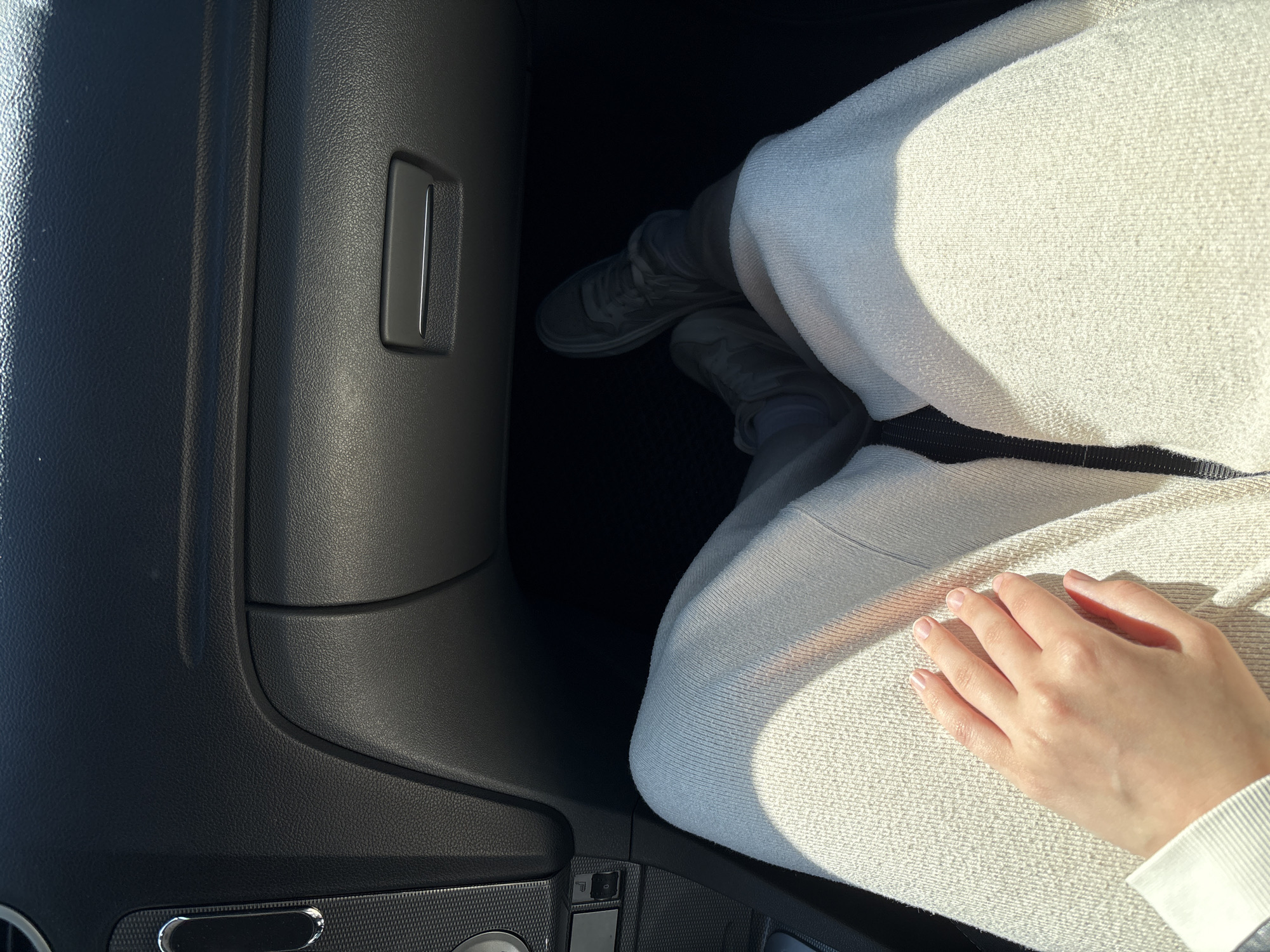 The image shows a close-up view of a person's legs and hand inside a car's passenger side. The person is wearing light-colored sweatpants and dark sneakers. A hand with manicured fingernails rests on the thigh. The car's interior features a black dashboard with a glove compartment and some controls on the left side. The environment suggests a sunny day, with light filtering into the car and casting shadows. The location is not specified in the image and cannot be determined.