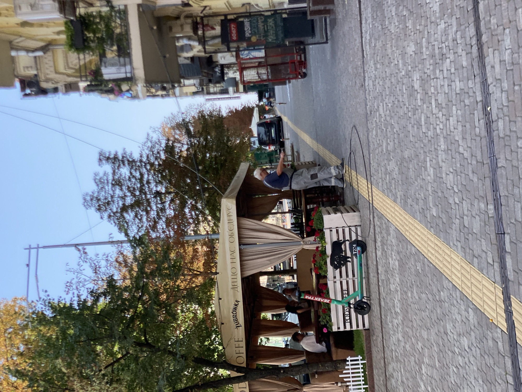 A man in grey overalls and a blue t-shirt is standing on a cobblestone street, holding a black hose connected to a white device. He appears to be cleaning or watering an area near a covered outdoor seating area of a cafe. The cafe has a large beige canopy supported by poles, with beige curtains drawn around the sides. Several people are seated at tables under the canopy. In front of the seating area is a green and white electric scooter with the word "ZELECTPA" written on it in red. Beside the scooter is a wooden planter box decorated with a silhouette of a buffalo. Trees with autumn foliage are visible behind the cafe. To the right of the street, a multi-story building has balconies with plants and air conditioning units. Signs with Cyrillic writing are visible on the building. The street is paved with grey cobblestones and has a yellow tactile paving strip along the sidewalk.
