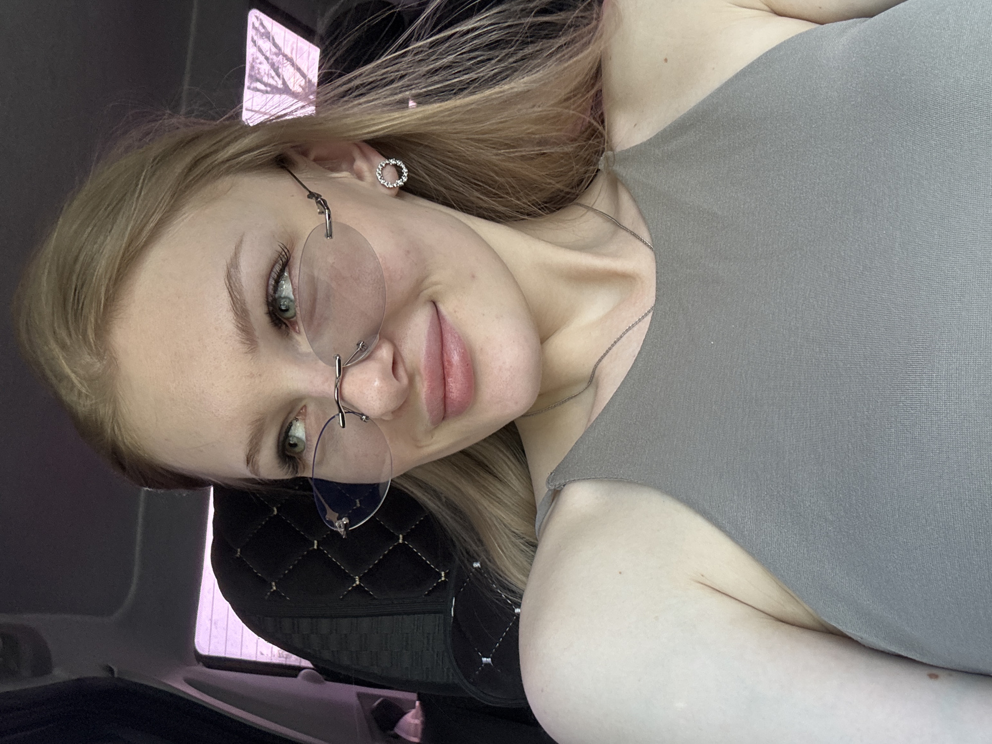 A fair-skinned young woman with blonde hair and light green eyes is posing in a car. She is wearing wire-rimmed glasses and a gray tank top. She also has a silver necklace and earrings. The car has black quilted headrests. Based on window reflections, the time of year appears to be winter. The location is Perm, Russia, according to information related to the image.