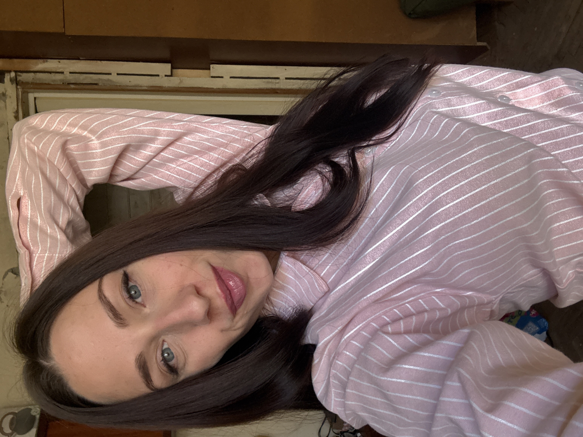 A selfie-style portrait captures a woman with long, dark brown hair and light blue-green eyes, wearing a light pink, striped long-sleeved shirt. Her hair flows down her left shoulder, and her gaze is directed towards the camera with a soft, slightly smiling expression, her lips tinted with light pink lipstick. The image is rotated 90 degrees counter-clockwise, suggesting a comfortable, informal pose.
The background reveals an indoor setting that appears rustic or in need of renovation. On the left side of the frame (when viewed upright), there's an unfinished wooden door or window frame with exposed wood and horizontal slats above it. The upper right portion of the background shows a dark wooden surface, possibly a shelf or beam, and a glimpse of a dark green object. The overall lighting is soft and ambient, suggesting an indoor, possibly daytime, environment. The woman's attire and the informal setting contribute to a casual atmosphere. There is no clearly discernible text in the image. The context provided indicates the location is Saint Petersburg, Russia.