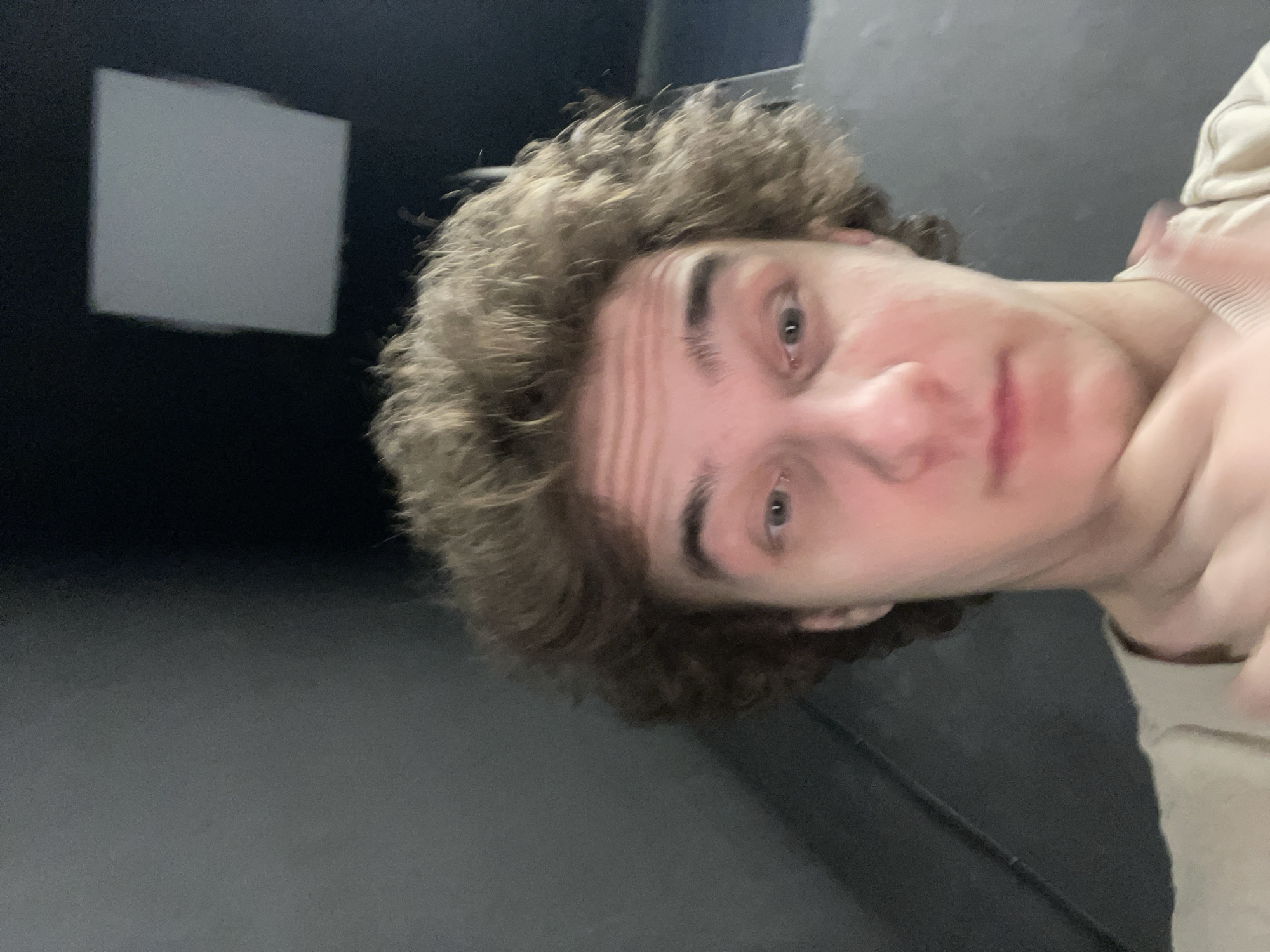 A low-angle selfie depicts a person with curly brown hair and fair skin. The individual's face is the primary subject, featuring gray eyes, a straight nose, and a neutral expression. The person is wearing a light-colored garment, possibly a sweatshirt. The background is dominated by dark gray surfaces, suggesting an indoor setting. In the upper left corner, a rectangular white shape is visible on a dark wall. There is no evidence in this image to support the location being in the city of Krasnoyarsk, Russia.