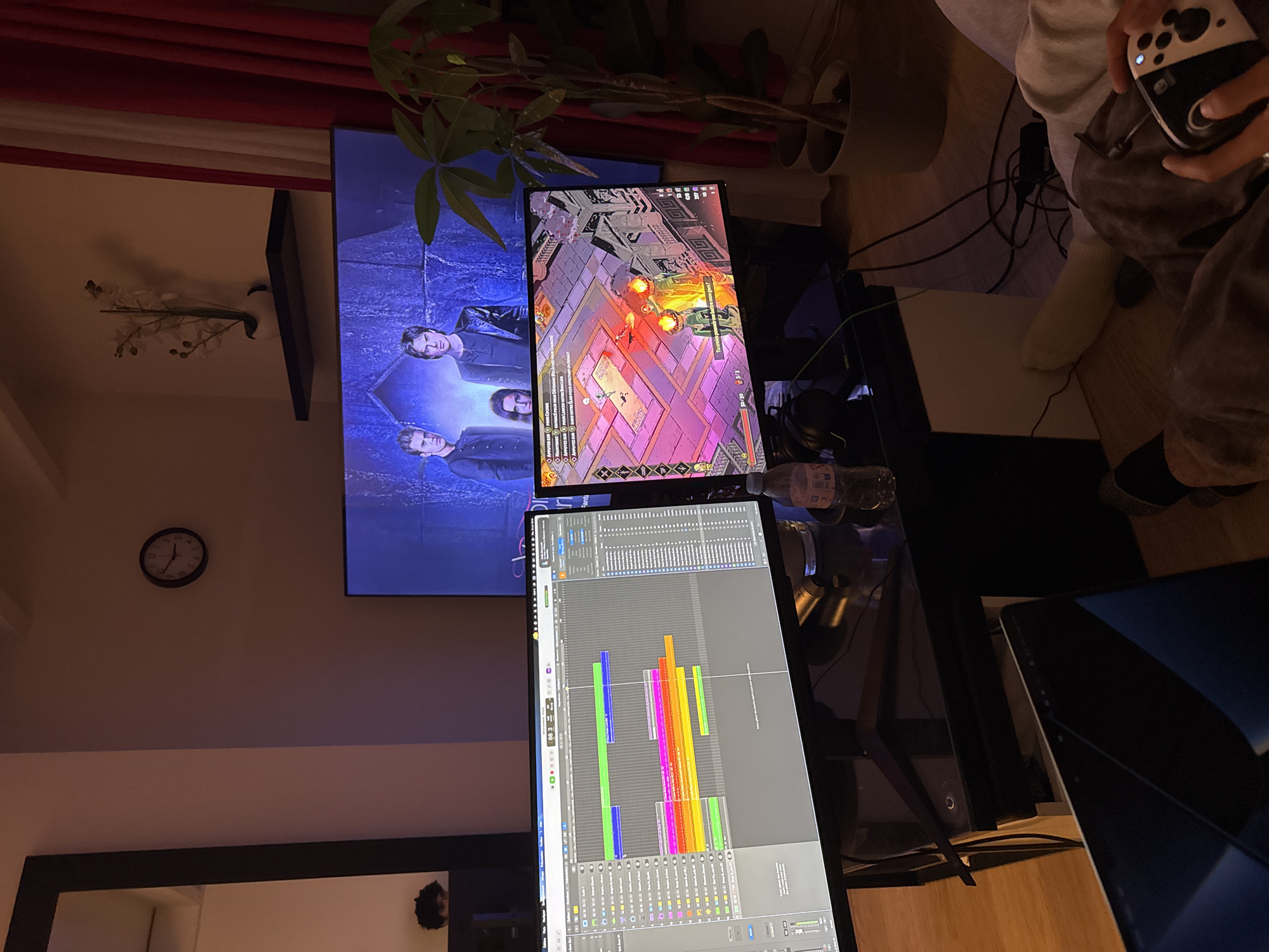 The image depicts a room with several monitors displaying various content. A large TV on the wall shows an image of four people, likely from a movie or TV show. Above the TV, a plant is visible. A smaller monitor displays a video game interface, and another shows a digital audio workstation (DAW) timeline with colorful tracks. A person is holding a game controller. A clock is visible on the wall, and a mirror can be seen reflecting part of the room. It is not possible to verify if this image contains the city of Paris.