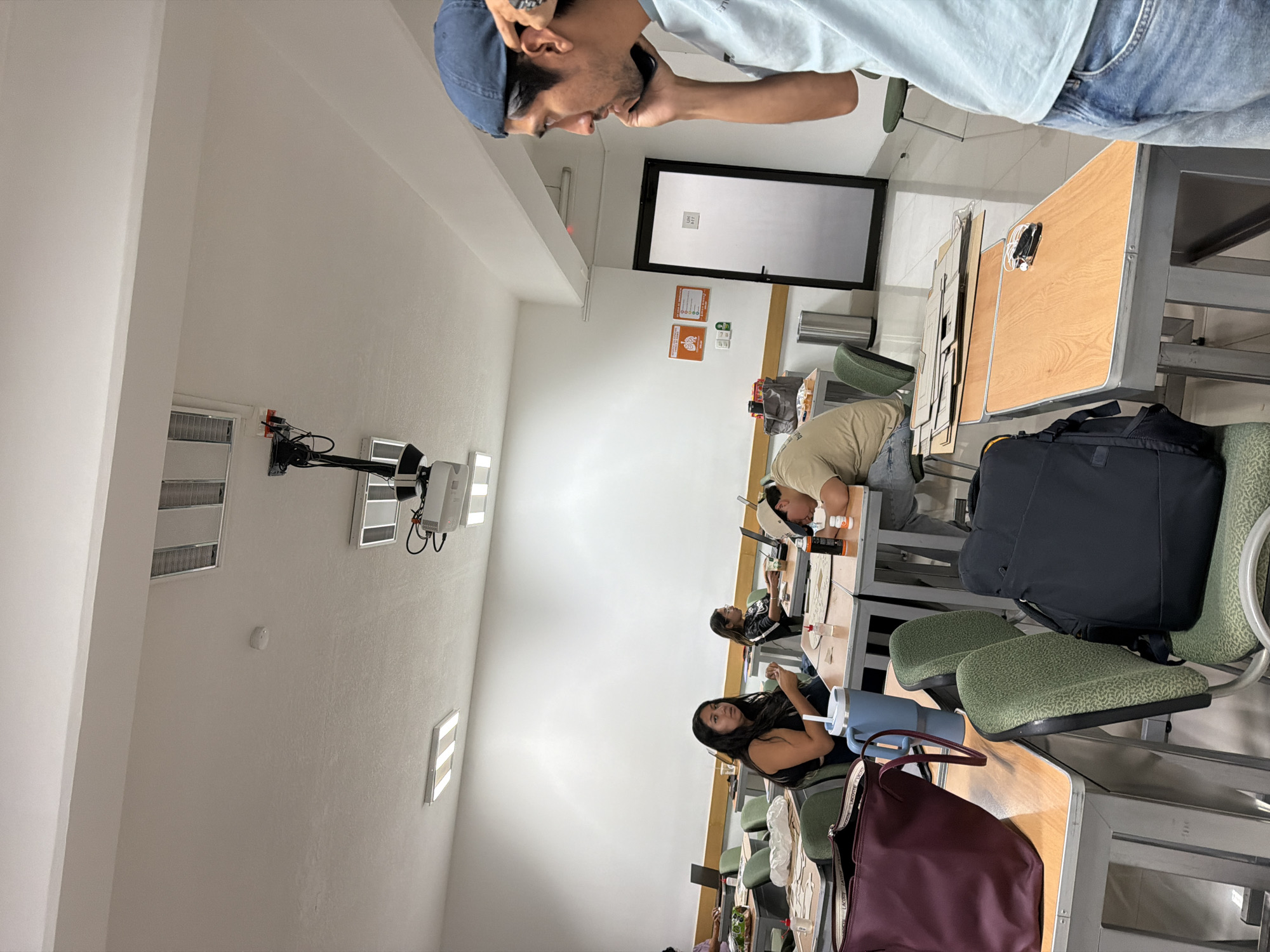 A classroom scene in San Andrés Cholula, Mexico. There are at least four students seated at desks in rows. A man wearing a blue cap and light blue shirt is on a mobile phone. The room has white walls and tiled flooring, with a whiteboard at the front of the room and a projector mounted on the ceiling. Individual desk chairs feature green fabric. Some students are interacting with objects on their desks.