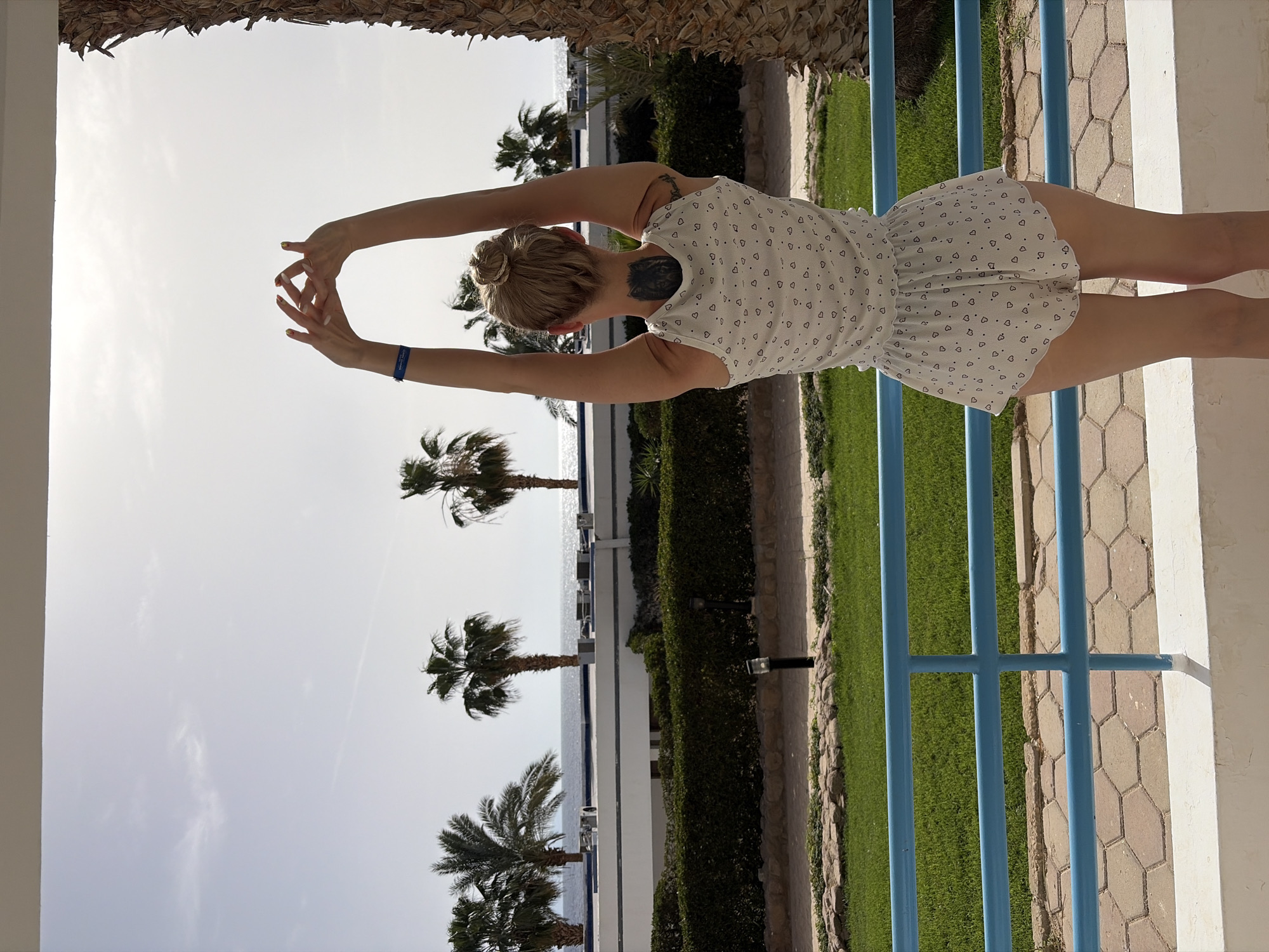 A woman stands facing away from the camera, stretching her arms overhead, near a blue railing and palm trees. The location is Sharm El Sheikh, Egypt, indicated by the tropical vegetation and coastal setting. The woman has a bun hairstyle and a tattoo on her neck. She wears a white dress with a small heart pattern. The scene includes a clear, bright sky, the sea, and manicured green lawns.