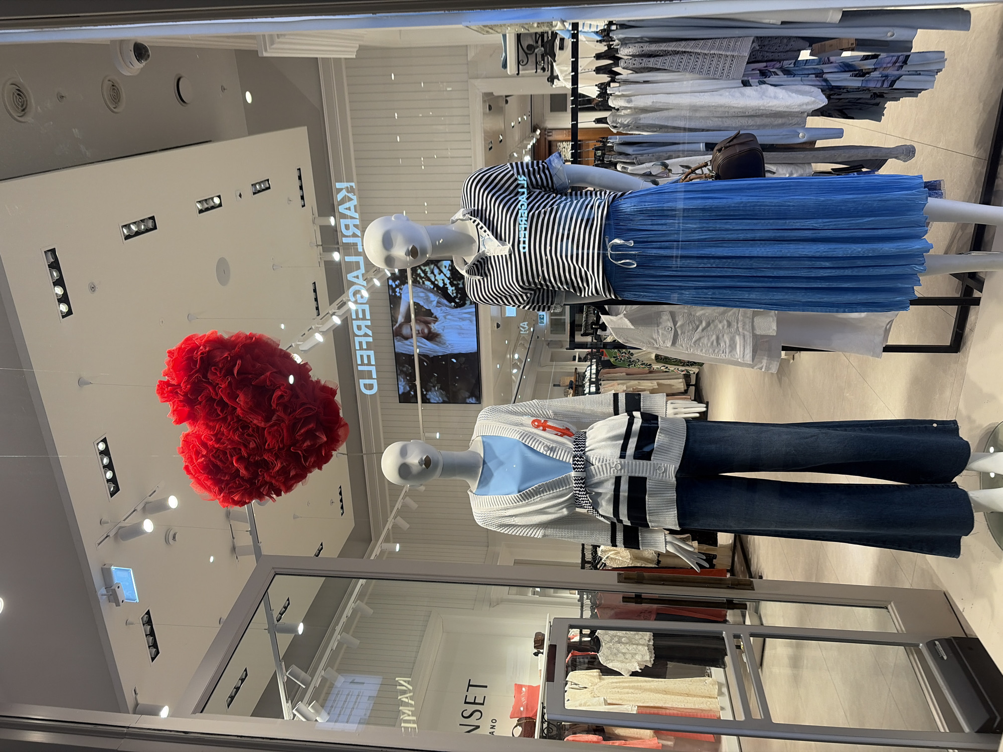 A storefront window display at a "KARL LAGERFELD" retail store in Saint Petersburg, Russia, is captured, with the image rotated 90 degrees clockwise. Two mannequins are the central subjects, showcasing clothing.
The upper mannequin wears a short-sleeved, black and white striped top with a ruffled collar and a pleated, light blue mid-length skirt. A small, dark brown handbag is visible near its waist. The lower mannequin is dressed in a light grey cardigan with dark stripes and a red anchor motif, worn over a light blue top, paired with dark blue, wide-leg jeans. Between the mannequins, a large, fluffy red heart decoration, made of ruffled fabric, hangs from the ceiling.
The store's interior is brightly illuminated by numerous recessed and track lights on the white ceiling. The brand name, "KARL LAGERFELD," is displayed in large, illuminated letters on an interior wall, appearing reversed due to the reflection off the glass. Another partially reflected name, "MANSET," is visible on a lower display shelf. Racks of clothing are visible in the background, and a large screen shows an image of a person, possibly a model. Reflections on the glass also include a security camera and ceiling vents. The setting suggests an indoor shopping environment, such as a mall, with the display designed to attract customers, possibly for a themed event given the prominent red heart.
