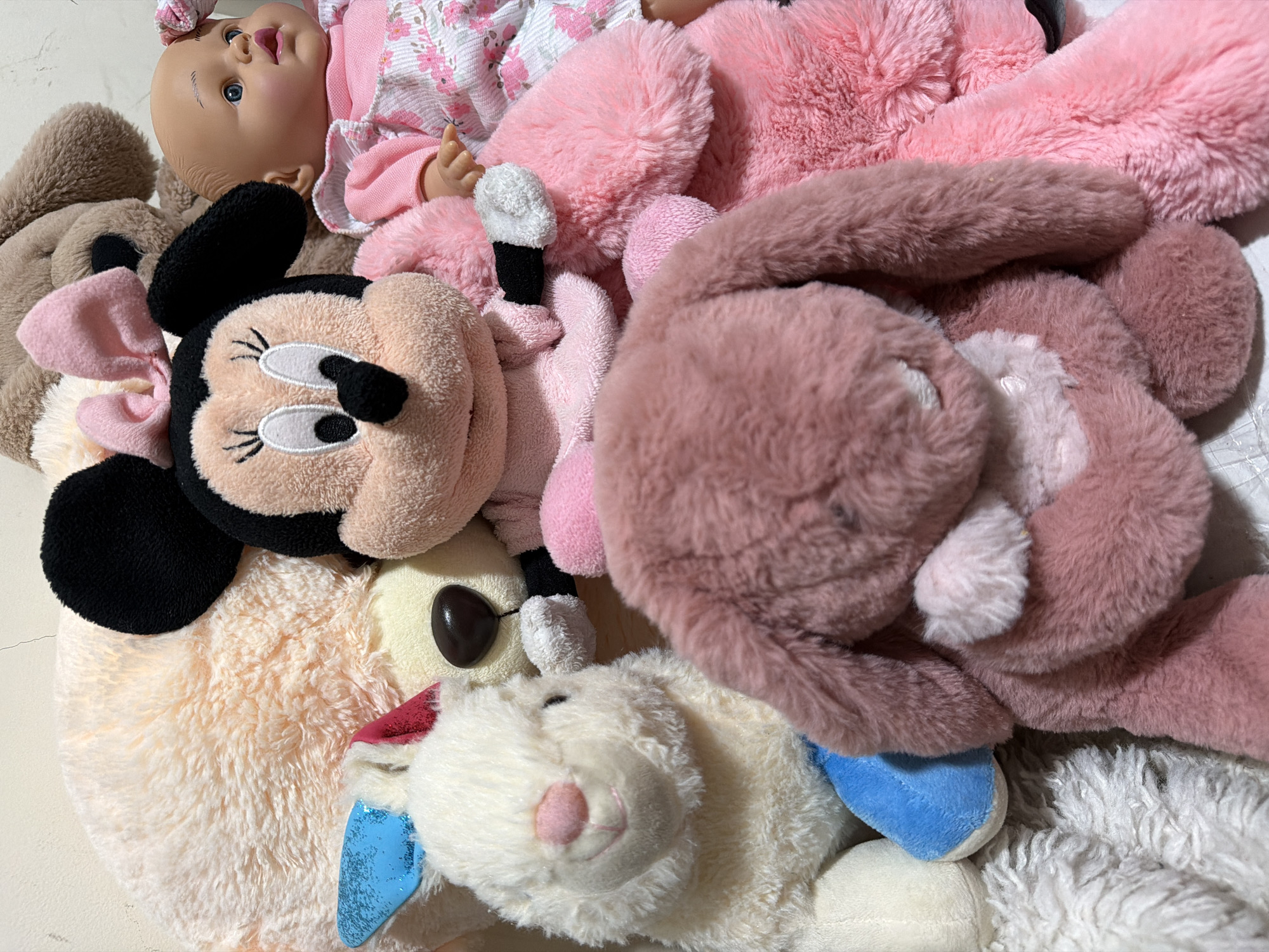 A close-up, overhead shot displays a collection of soft, plush stuffed toys. In the foreground, a pink fuzzy bunny is positioned on its back, with its head slightly turned to the left. To its left, a white bunny with blue and pink accents is seated upright, facing forward. Behind the white bunny and to the right of the pink bunny, a plush Minnie Mouse toy is centrally located, its head turned towards the viewer. To the right of Minnie Mouse, a baby doll with blue eyes, a pink bow, and a pink floral dress is visible. In the background, a large, fuzzy tan stuffed animal and a light pink stuffed animal are partially visible. The toys are arranged on a flat surface, appearing to be a bed or floor. The lighting is soft and diffused, suggesting an indoor setting. The image does not contain any information or context that would allow for the determination of its location in Central Jakarta, Indonesia.