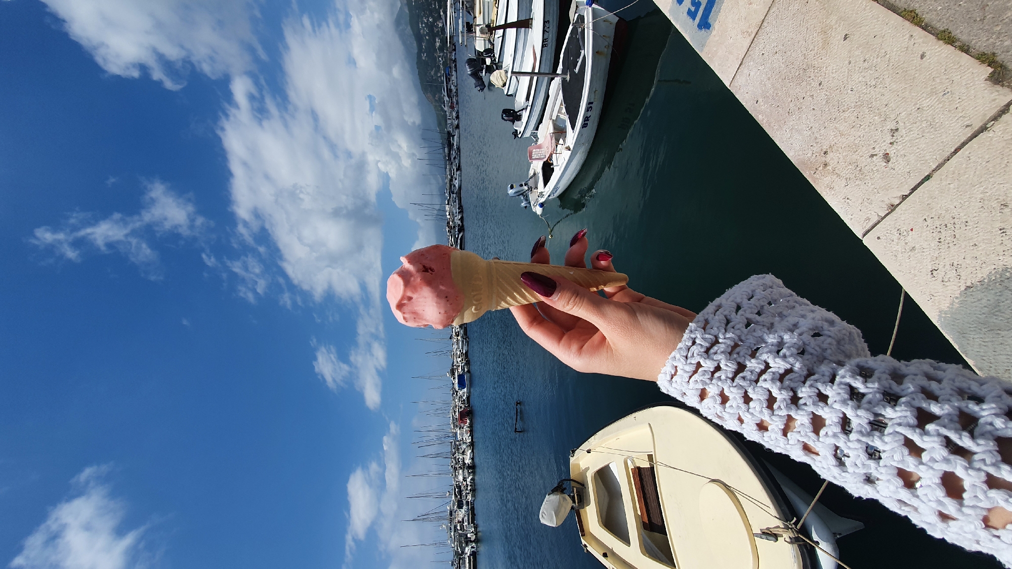 Here is a description of the image:
The image, taken in the city of Bar, Montenegro, shows a hand holding an ice cream cone against a backdrop of a marina filled with boats. The ice cream is pink and appears to be strawberry flavor. The hand is wearing a crocheted white sleeve. The marina is filled with numerous boats of varying sizes, with a clear blue sky with puffy white clouds above. The water in the marina is a dark blue-green color. A concrete pier or dock is visible in the foreground.