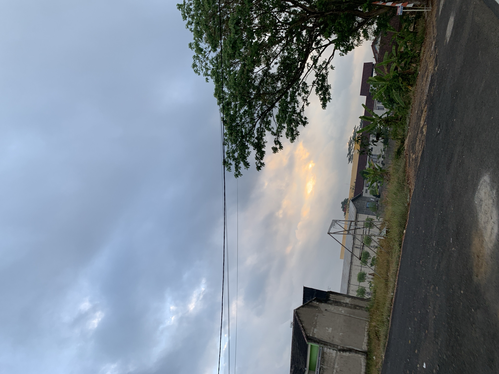 The image captures a view of a street in Cijeungjing, Indonesia, under a cloudy sky with hints of sunset. In the foreground, a dark asphalt road with some debris is visible. To the left, a partially constructed building with concrete walls and a tiled roof stands. Further back, there are several buildings with white and brown facades, some with red roofs, surrounded by greenery. A large tree with dense foliage occupies the right side of the frame, its branches extending towards the sky. Power lines are strung across the upper portion of the image. The scene appears to be a residential or semi-urban area during the late afternoon or early evening.