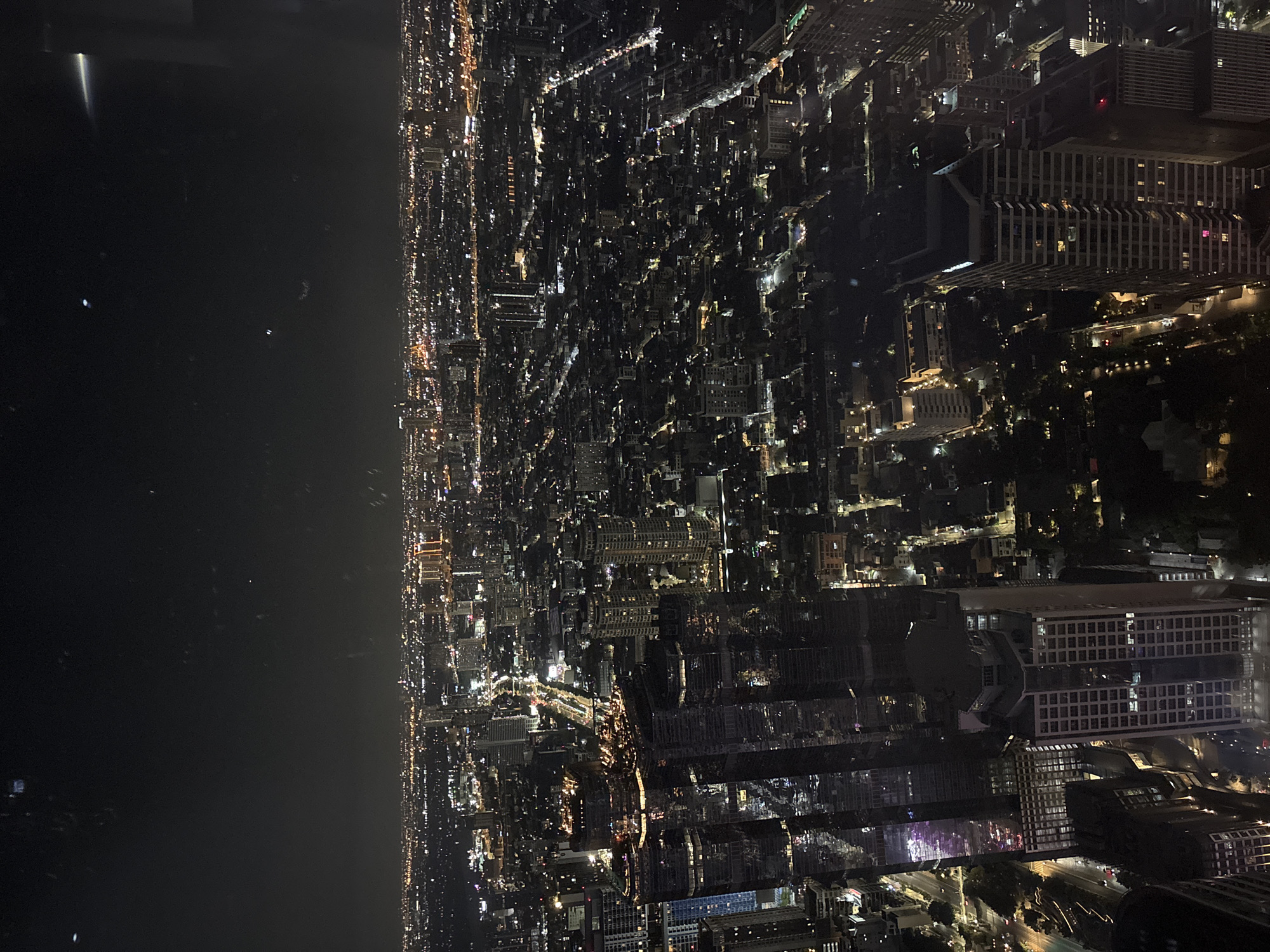 This media file presents an expansive aerial view of Bangkok, Thailand, captured at night from a high vantage point. The scene is dominated by a sprawling urban landscape, densely packed with thousands of illuminated buildings, streets, and structures. The artificial lights from the city create a glittering, intricate pattern that stretches across the entire frame, showcasing the vastness of the metropolis.
To the left side of the image, a large, dark expanse of water is visible, which appears to be the Chao Phraya River. A few scattered lights are reflected on or emerge from the water, suggesting distant shores or water traffic. The sky above the cityscape and the river is dark and clear, indicating good visibility without heavy clouds.
The image conveys the vibrant energy and continuous activity of a major city after dark, characterized by its pervasive artificial illumination. While specific human figures or vehicles are not individually discernible due to the high altitude, the sheer volume of lights implies a bustling urban environment. The architecture of the visible buildings, particularly in the cropped sections, appears modern and varied, consistent with a contemporary Asian capital. No visible text is present within the image.