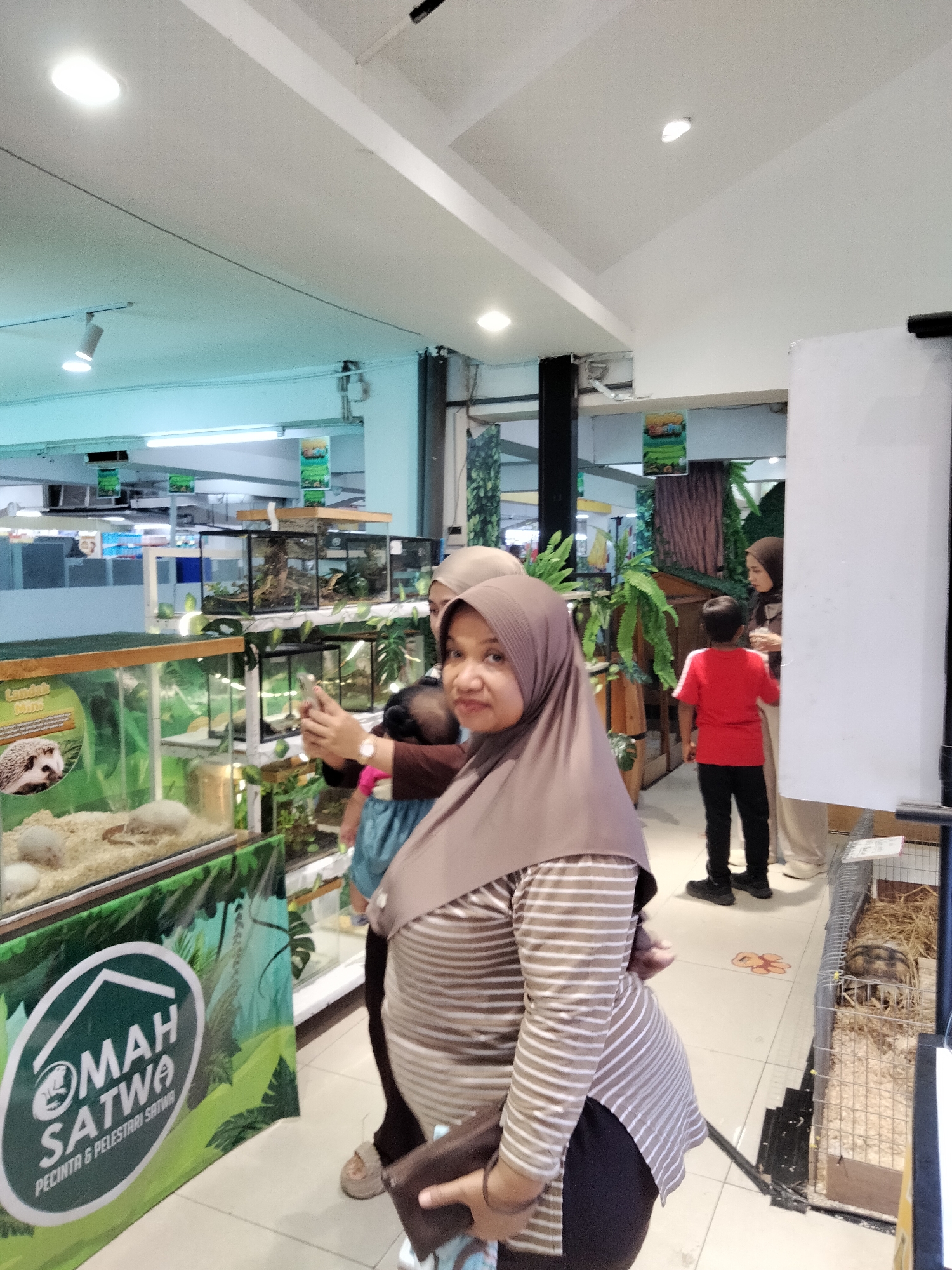 The image depicts an indoor scene, likely a pet store or animal exhibition, with several people and enclosures containing animals. In the foreground, a woman wearing a hijab and a striped shirt is holding a smartphone and appears to be taking a picture or recording a video. She is holding a dark-colored clutch purse in her other hand. A child is visible behind her, also wearing a hijab. To the right, a young boy in a red shirt and black pants stands near a woman wearing a cream-colored hijab and pants, who is looking at the boy. Further back, there are various glass enclosures with animals inside, including what appears to be a hedgehog in one of the foreground enclosures. The environment features bright overhead lighting and shelves displaying plants and animal habitats. A banner with text and graphics, including a house symbol and the words "OMAH SATWA PECINTA & PELESTARI SATWA," is prominently displayed on the left side of the image. The flooring is tiled. The overall impression is a public space dedicated to the display and appreciation of animals. The presence of the banner and the style of the signage suggest a location in Indonesia. While the specific location of "Kecamatan Pati, Indonesia" is provided as context, there are no explicit visual cues within the image itself that definitively confirm this precise geographical identification.